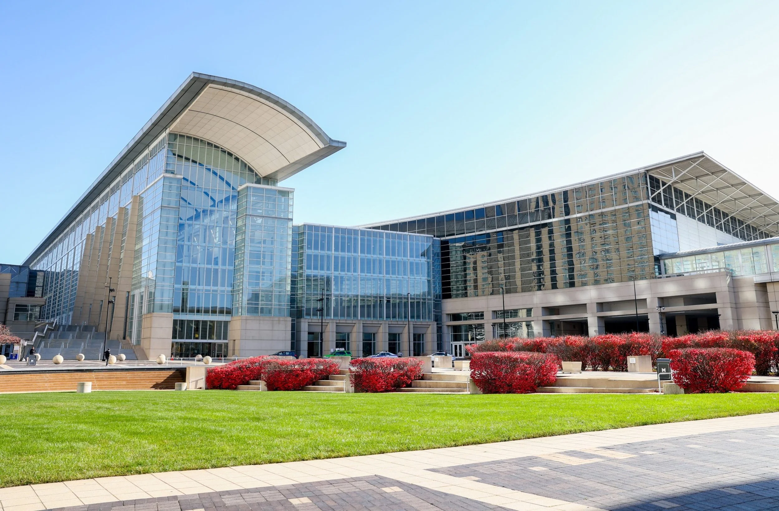 Modern glass building with a curved roof, reflecting the sky, surrounded by a lawn and red bushes.