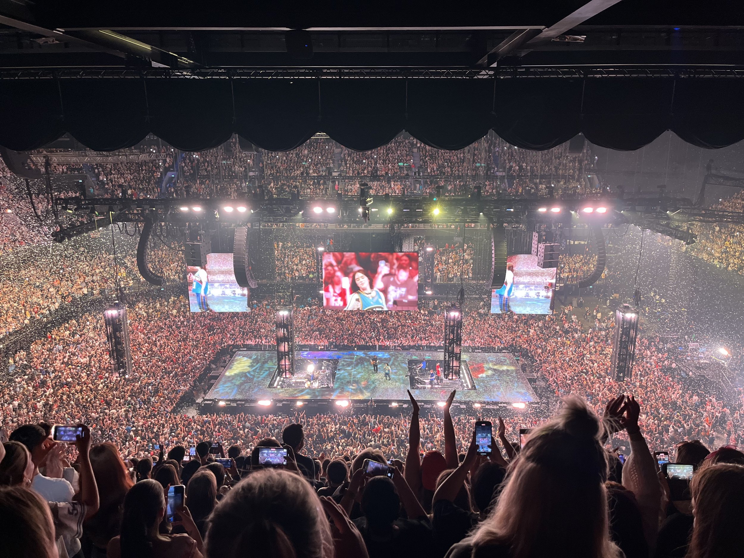A large indoor concert venue filled with a cheering crowd, stage lighting, large screens displaying performers, and confetti in the air.
