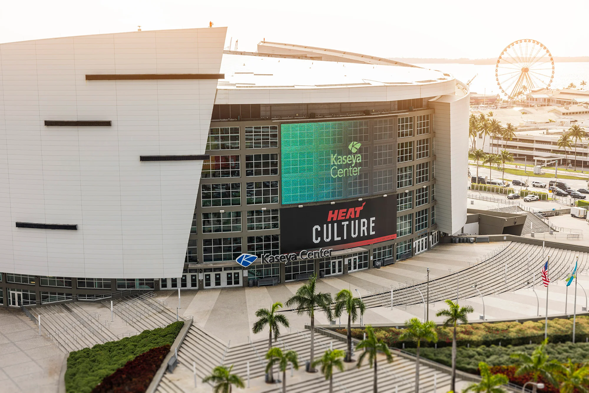 Exterior of the Kaseya Center with large digital screens displaying the event 'Heat Culture' and 'Kaseya Center' branding. Palm trees and flags are in the foreground, with a parking lot and a Ferris wheel in the background.