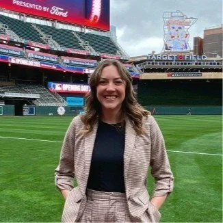 Woman smiling on a baseball field near the stands at Target Field, with a large scoreboard and person-shaped art display in the background.