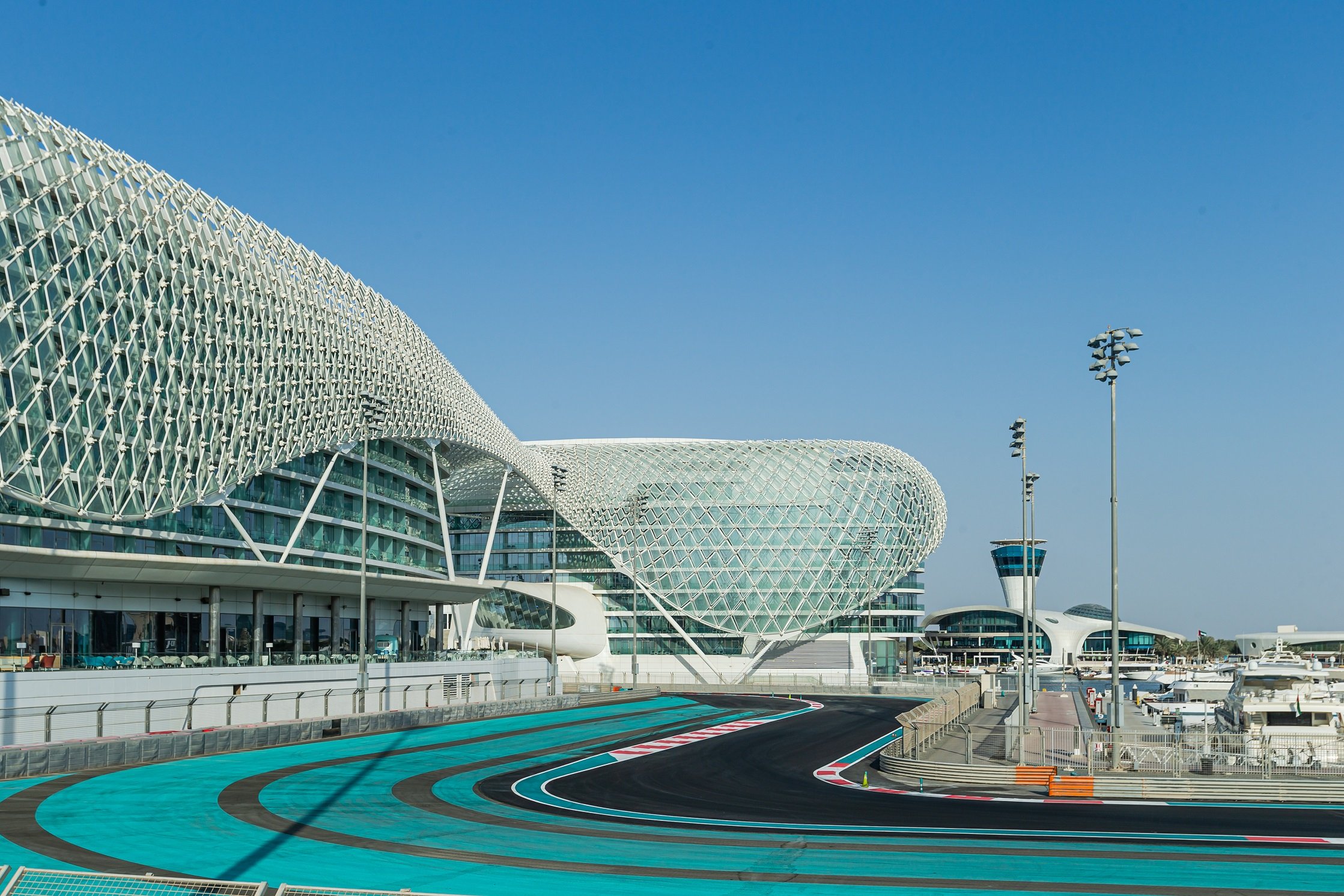 Modern airport terminal with a distinctive curved architectural design, situating an area with a racetrack and parked yachts, under a clear blue sky.