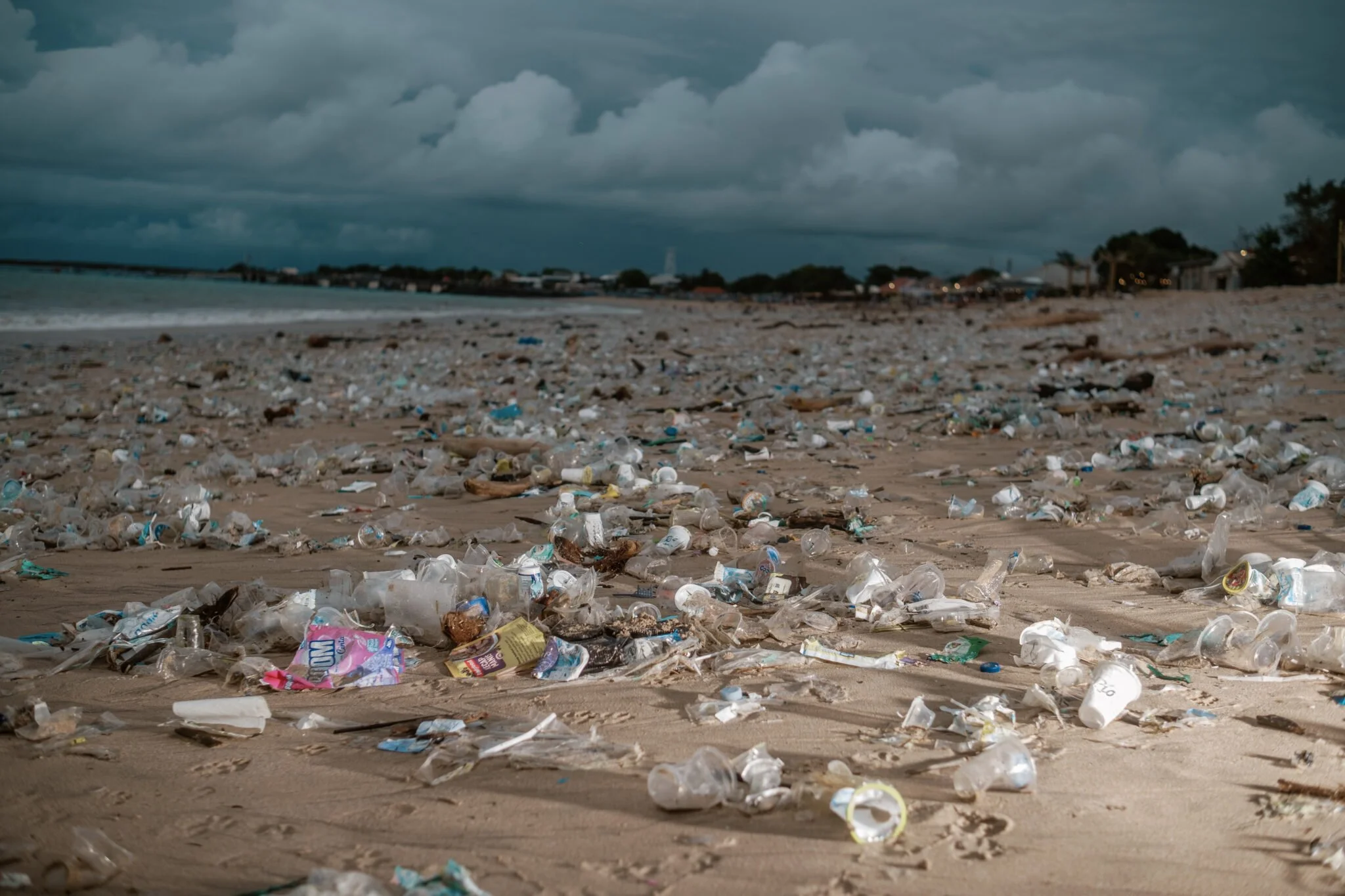 Beach covered with scattered plastic bottles, bags, and debris under a cloudy sky.
