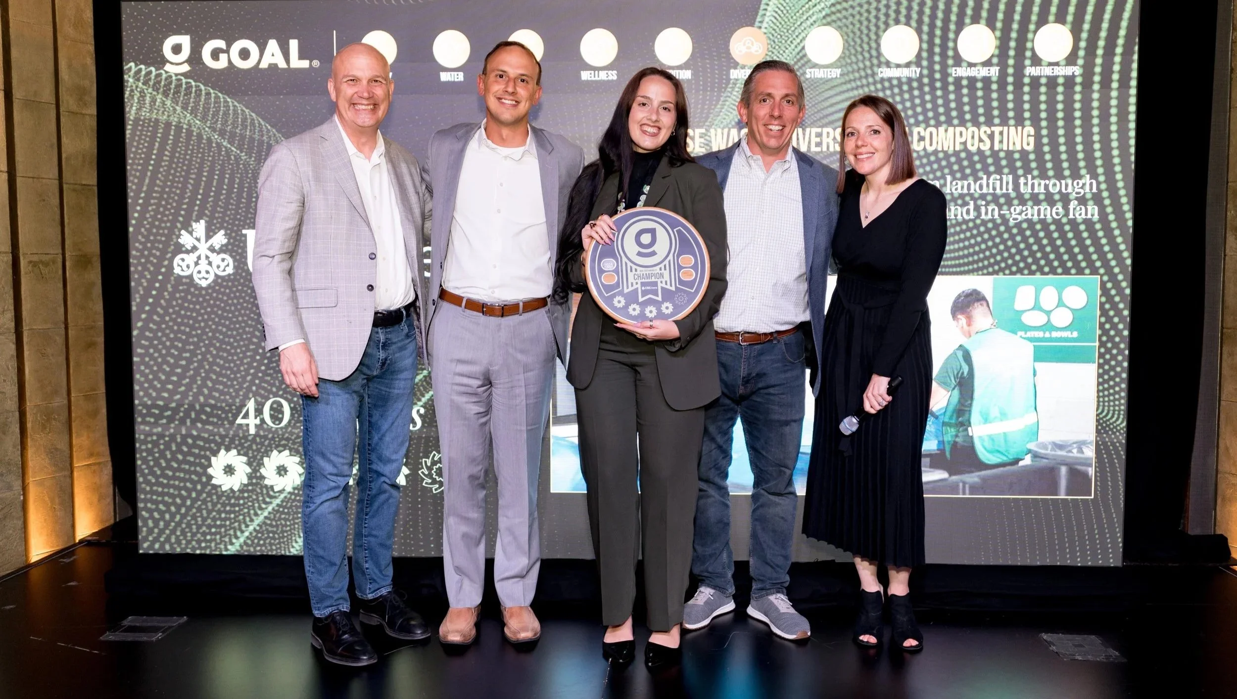 Six people standing on stage smiling, with a digital screen behind them displaying text and graphics, one woman in the center holding a round award with the letter 'g' on it.