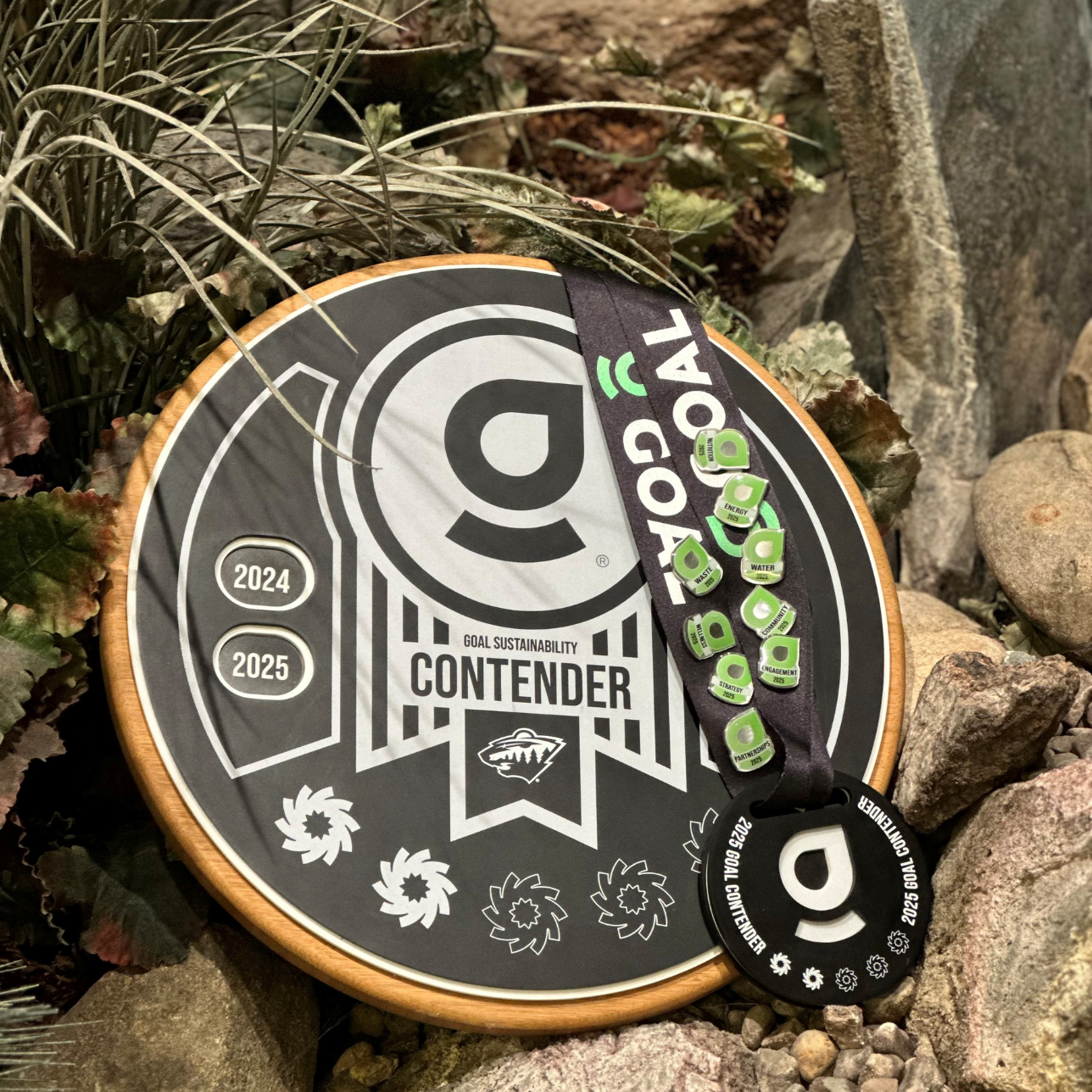 A black and white goal sustainability award plaque labeled "Contender" with the year range 2024-2025 and the logo of the Minnesota Wild hockey team featuring a wilderness scene. There is a round black medal attached to a lanyard with the word "D GOAL" and the years 2024-2025, along with small green and white pins displaying ecological icons like water drops, trees, and recycling symbols, placed on the plaque. The objects are on rocks with some plants around.