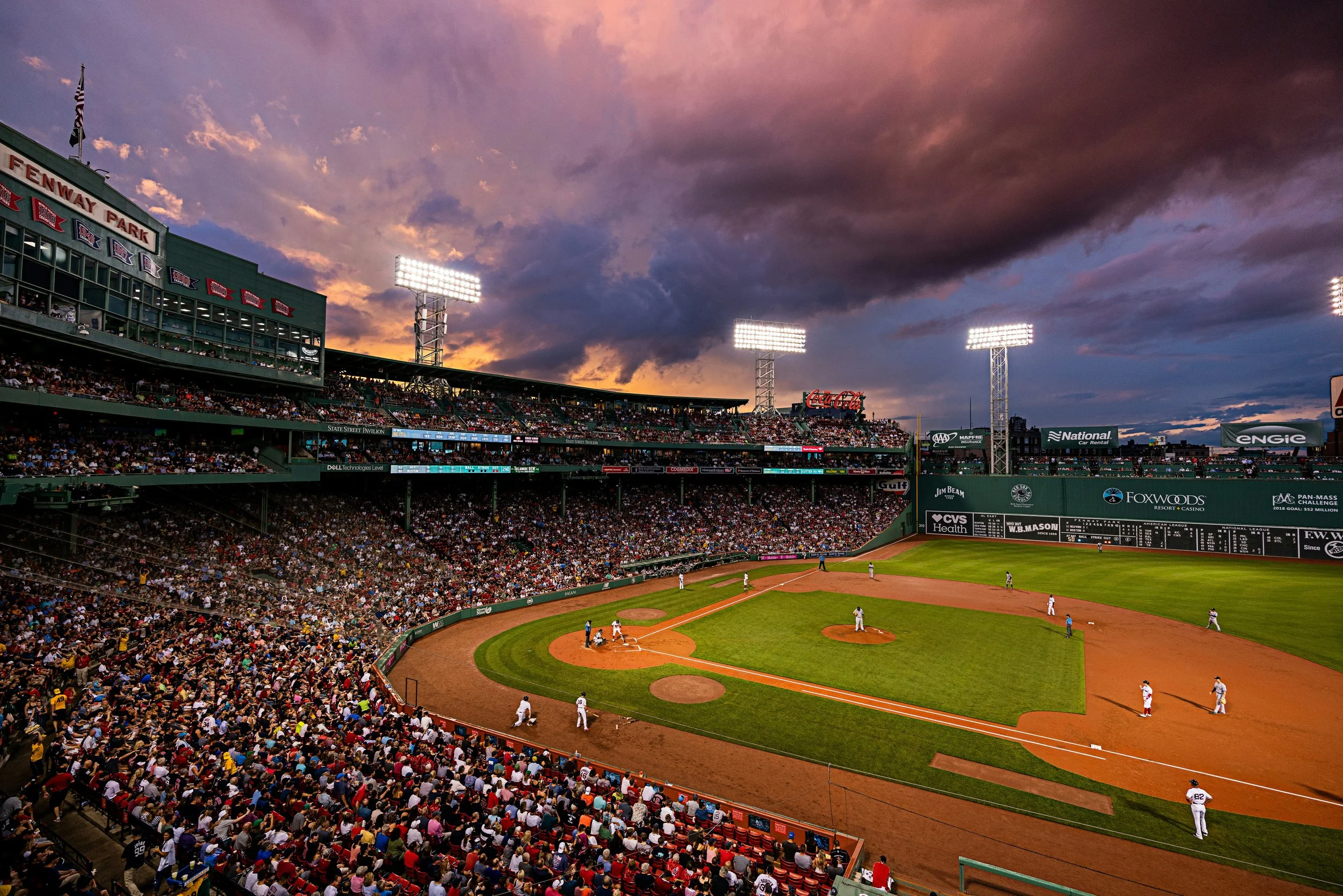 A baseball game in progress at Fenway Park during sunset, with players on the field and a crowd in the stands under a cloudy sky.