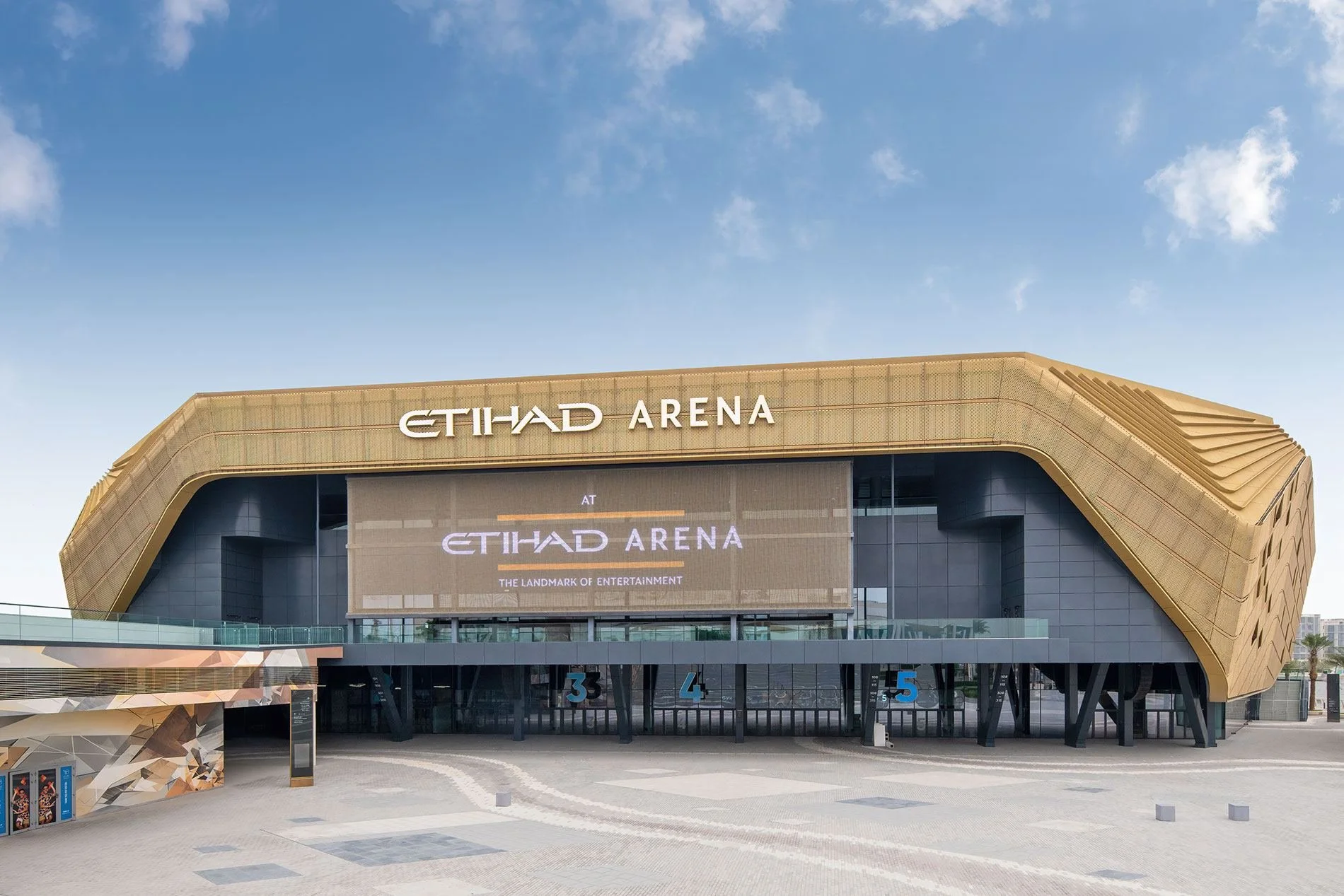 Exterior view of Etihad Arena with large digital screen displaying its name, a spacious plaza in front, and a partly cloudy sky above.
