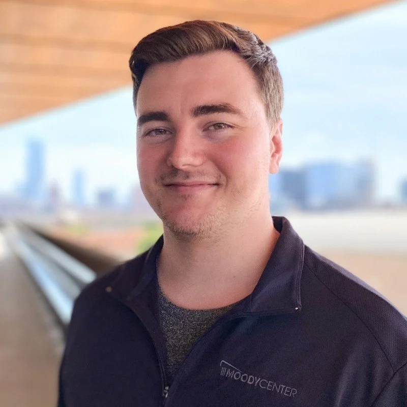 A young man with short brown hair, wearing a dark zip-up jacket with the Moody Center logo, smiling outdoors with a city skyline in the background.