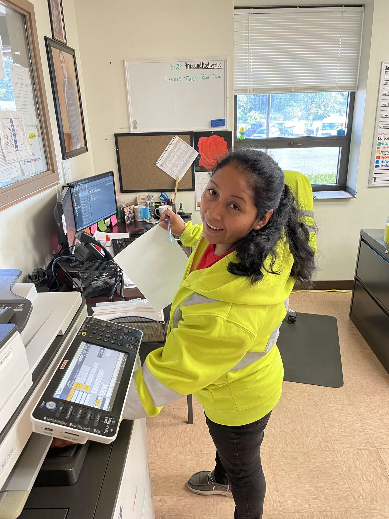 A woman in a yellow safety jacket standing at a print shop or office, holding a sheet of paper and smiling at the camera.