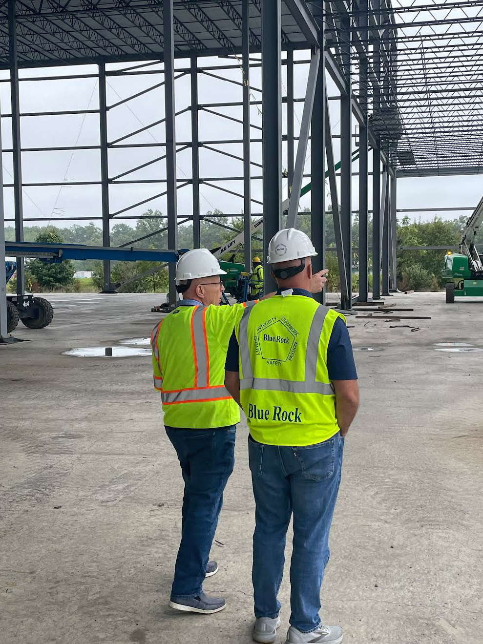 Two construction workers wearing safety vests and hard hats inspecting a building under construction with steel framework and construction equipment.