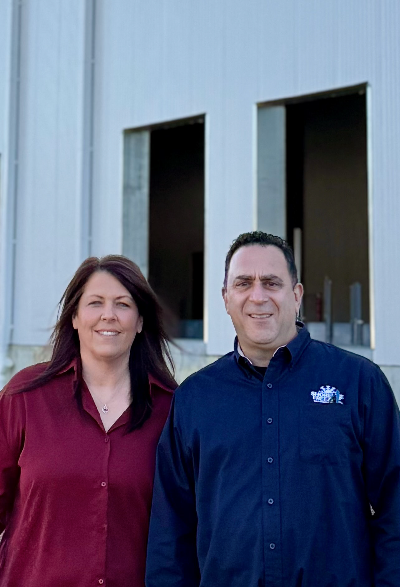 A woman with long dark hair wearing a red shirt standing next to a man with short dark hair wearing a navy blue button-up shirt with a Shoreline Freezers logo, in front of a cold storage warehouse with large open doorways.