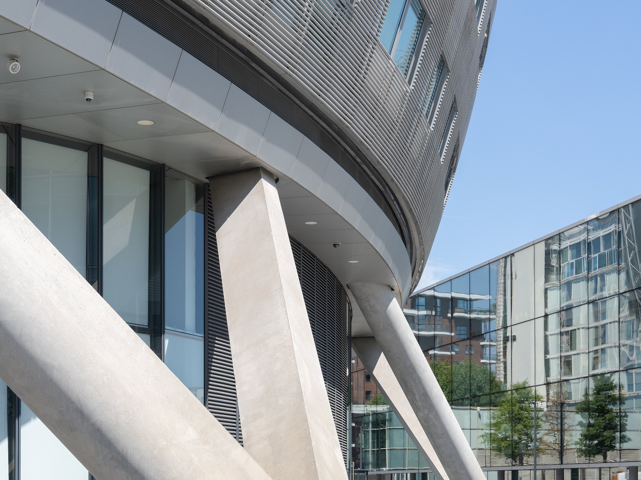 Modern commercial building with glass walls and angled concrete support columns, reflecting surrounding structures and trees under a clear blue sky.