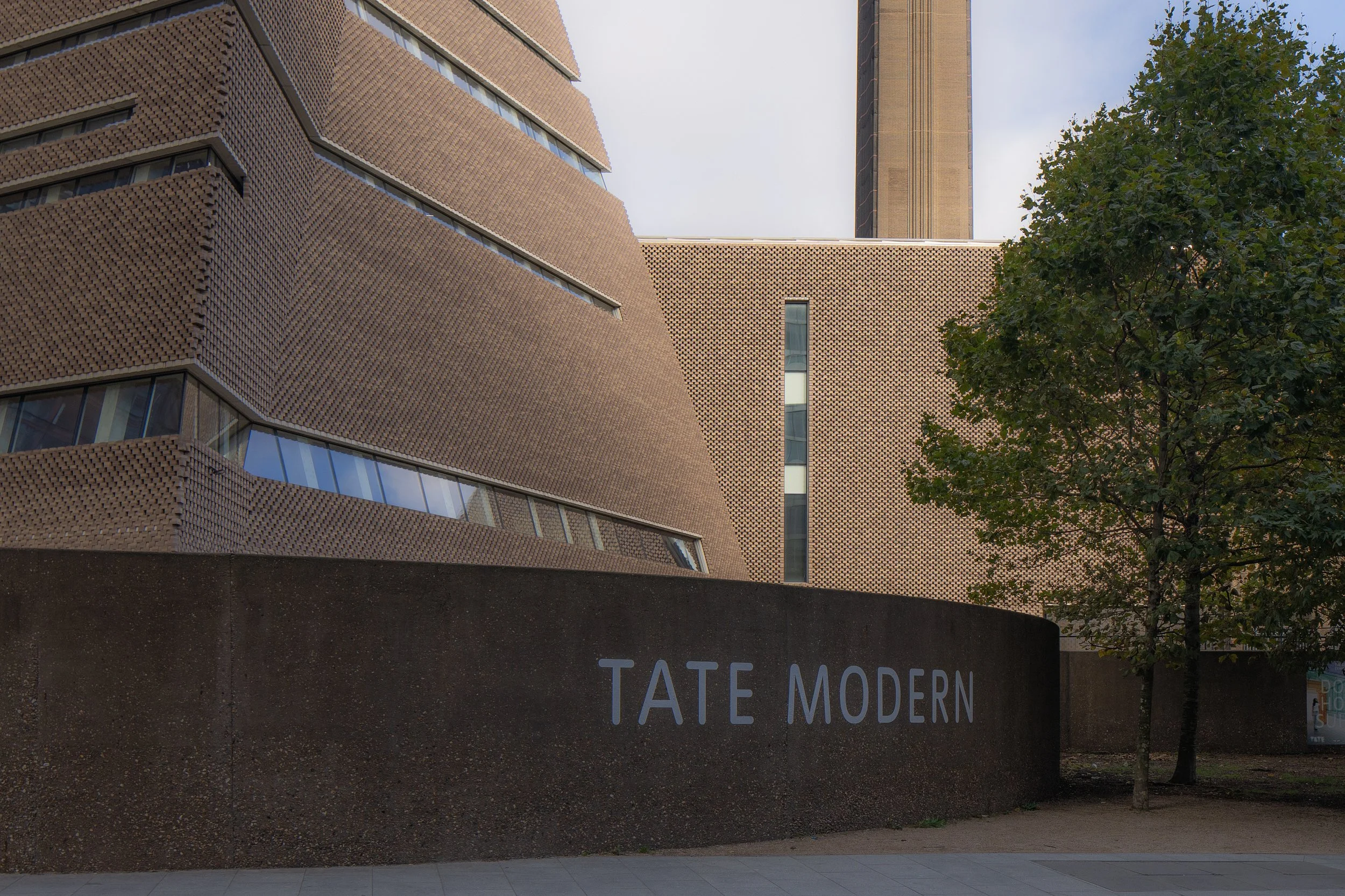 Photo of the Tate Modern building with its distinctive modern architecture, featuring textured brown brick walls, large windows, and the building's name on a concrete wall in front, alongside a tree.