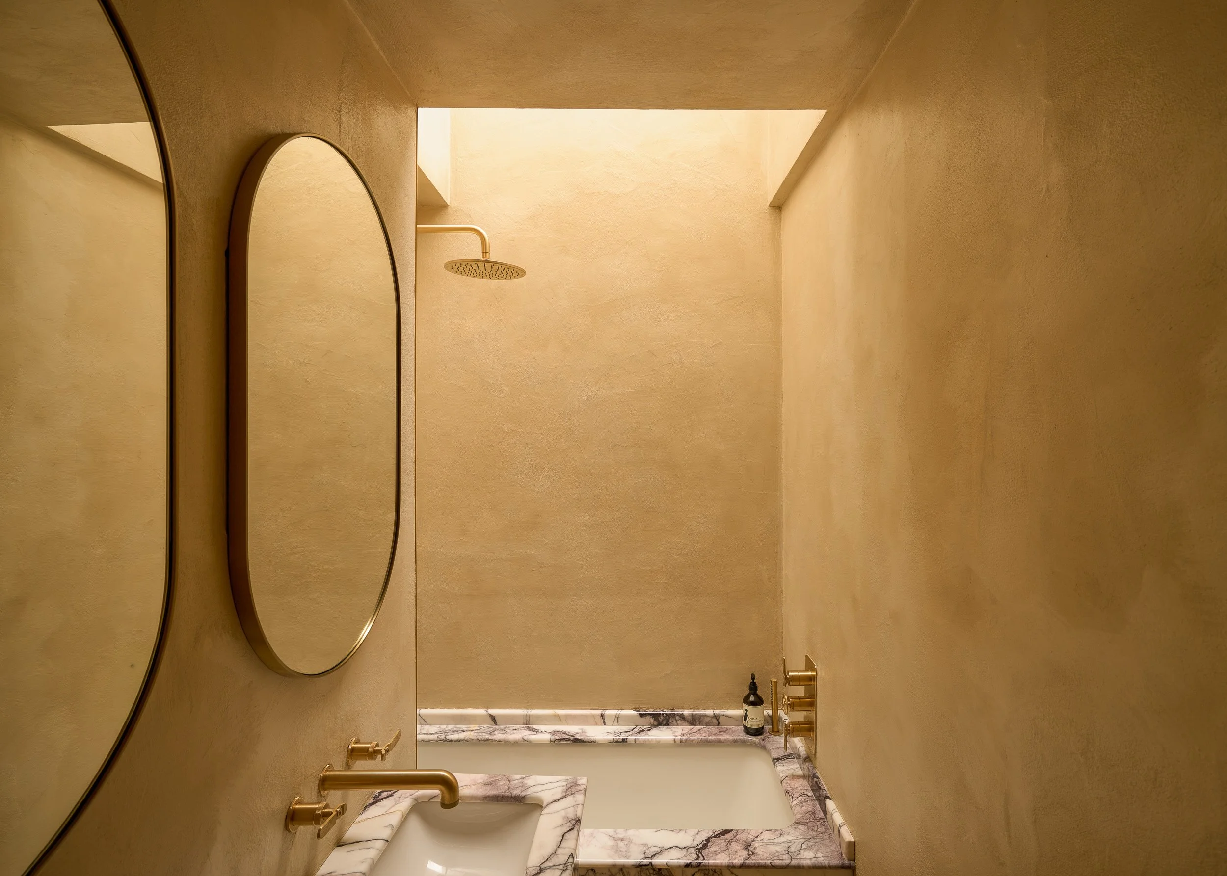 Minimalist bathroom with a marble countertop, oval mirrors, gold fixtures, a showerhead, and a small bottle of soap on the marble edge.