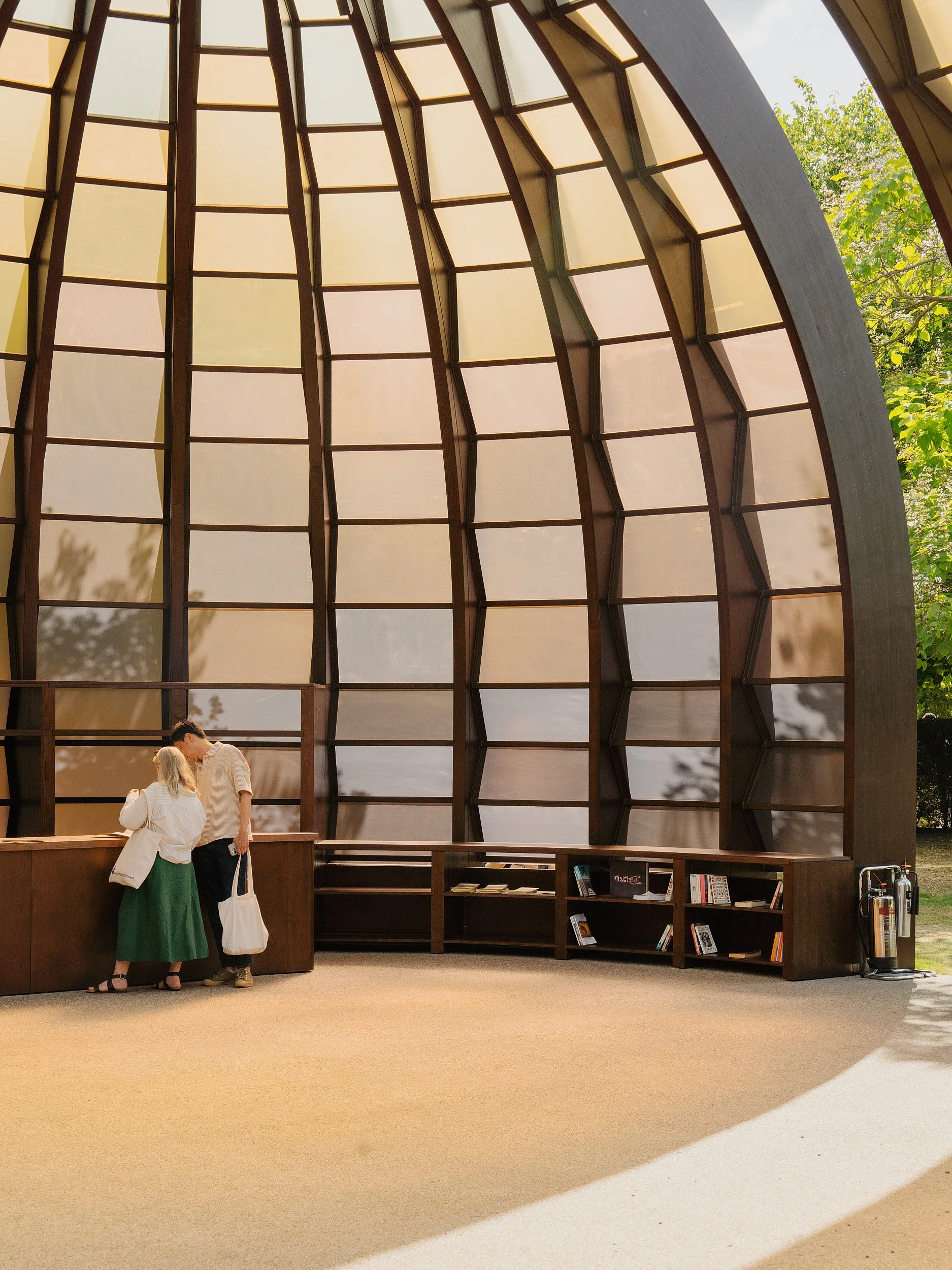 Two people at a wooden reception desk inside a building with large, curved, geometric windows allowing natural light to fill the space, with bookshelves and outdoor greenery visible.