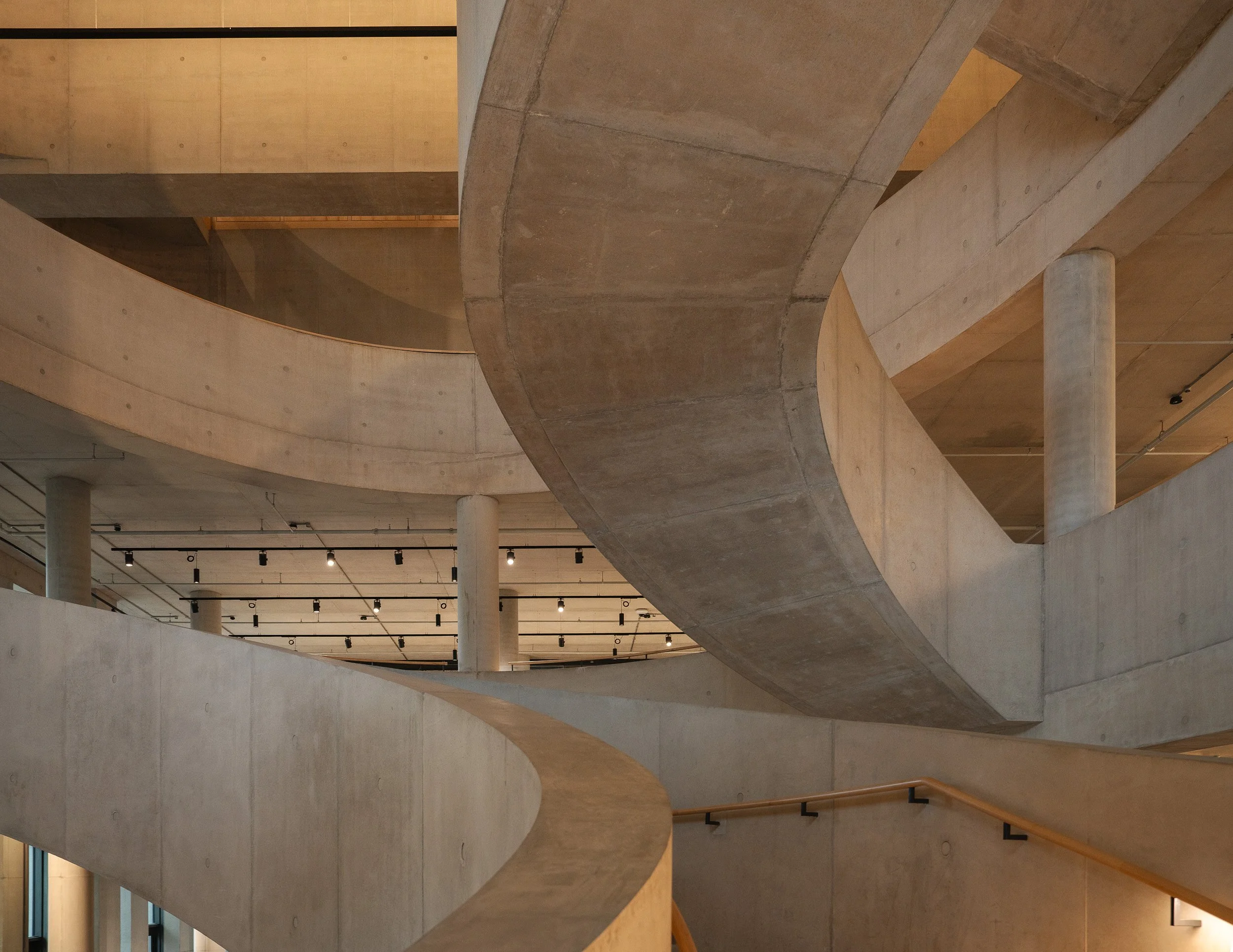 Interior view of a modern building with multiple winding concrete staircases and wooden railings, illuminated by ceiling lights.