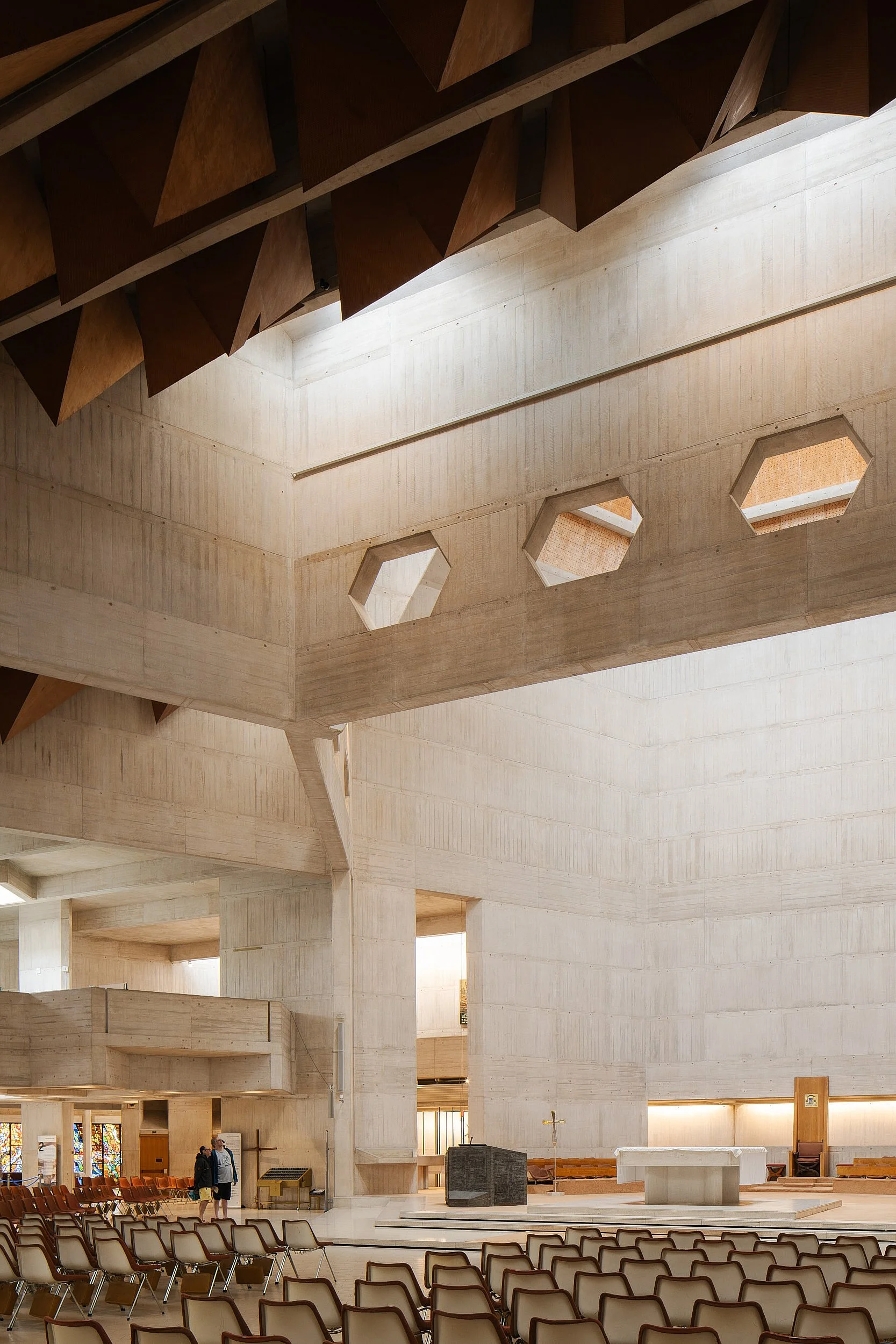 Interior of a church with modern architectural design, including a high ceiling with geometric wooden shapes, large hexagonal windows, and a spacious seating area with rows of chairs.