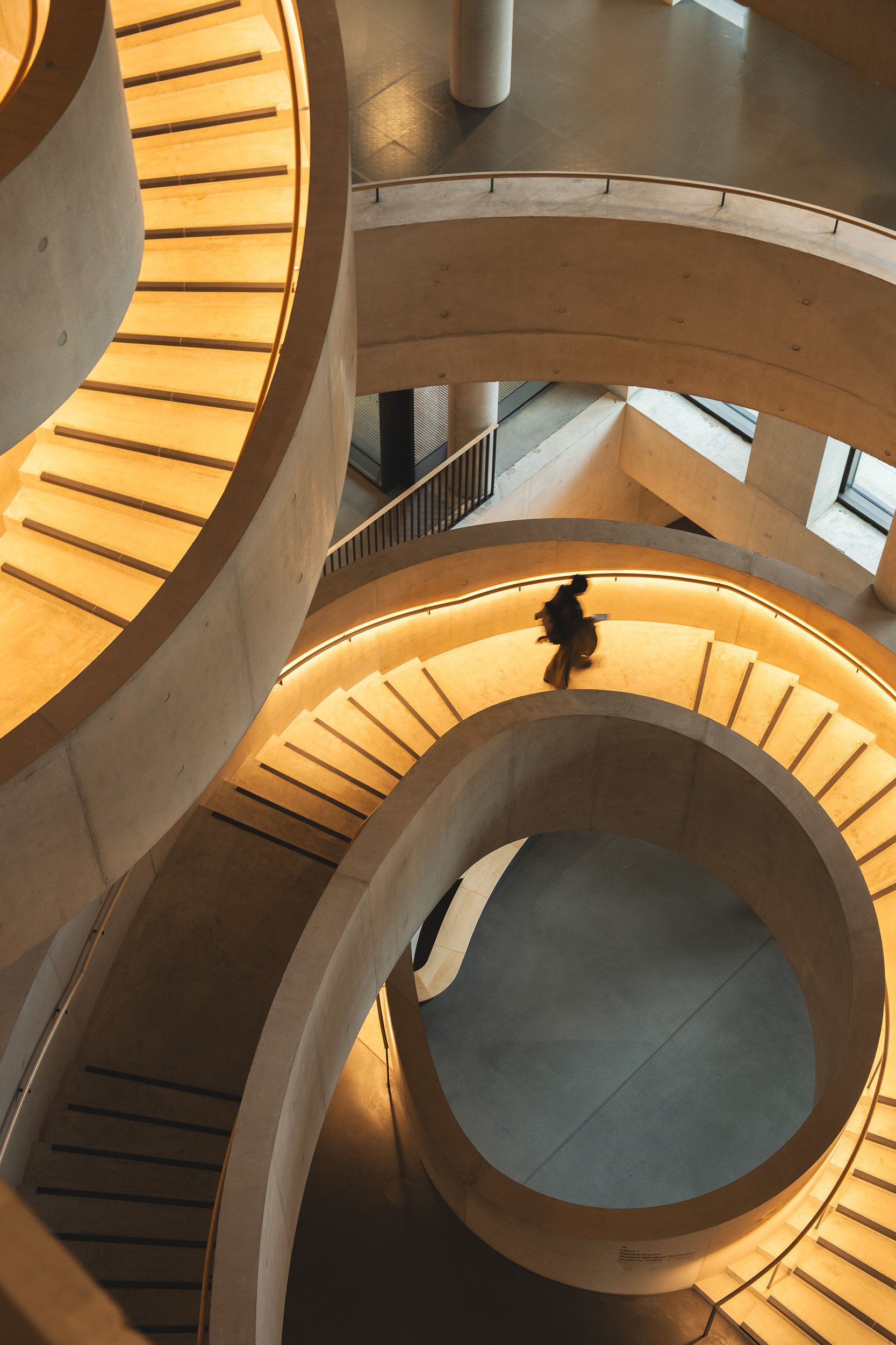 Interior view of a modern multi-level building showcasing a spiral staircase with illuminated yellow lighting, concrete surfaces, and a person walking inside.