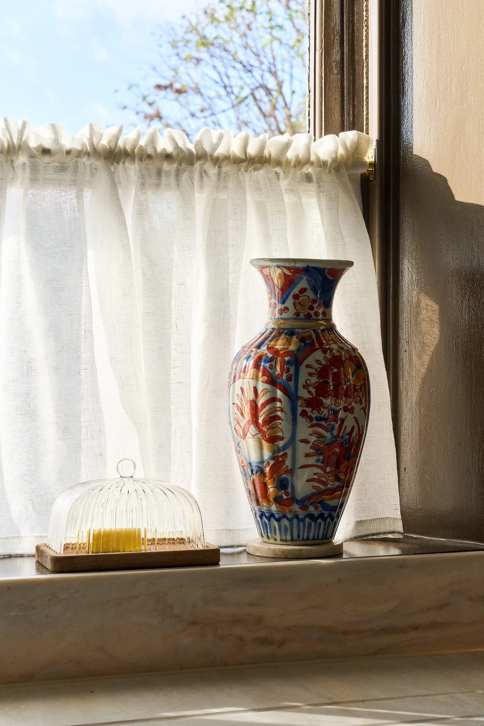 Decorative ceramic vase with colorful floral and bird pattern on windowsill, beside a small glass dish with candles, in front of a window with a white curtain and blue sky outside.