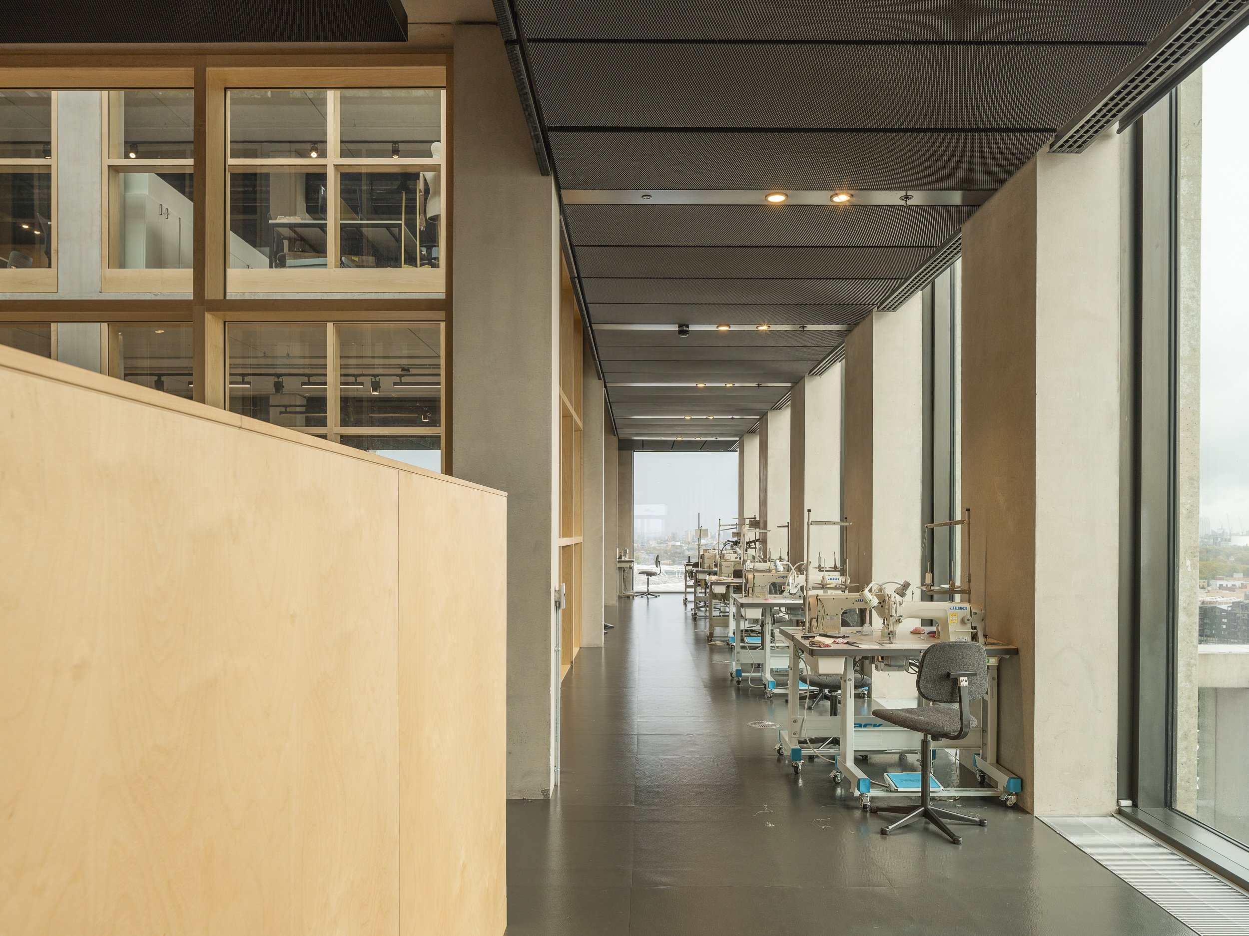 An industrial workspace with multiple sewing machines and chairs lined up along large windows, with a view of the city skyline outside.