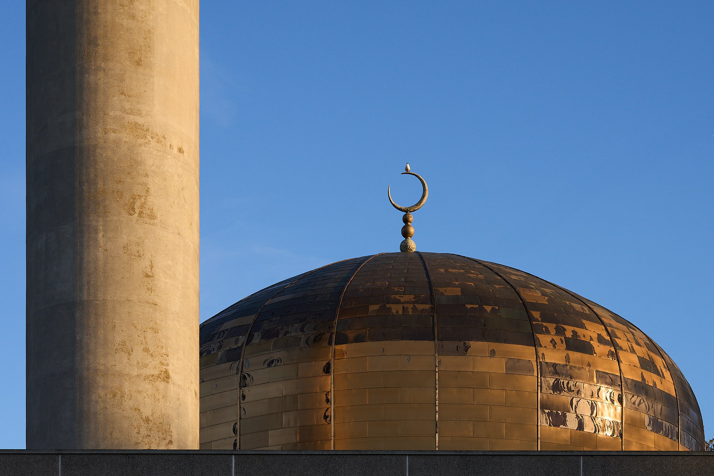Close-up of a mosque dome with a crescent moon and star finial, part of a minaret, against a clear blue sky.