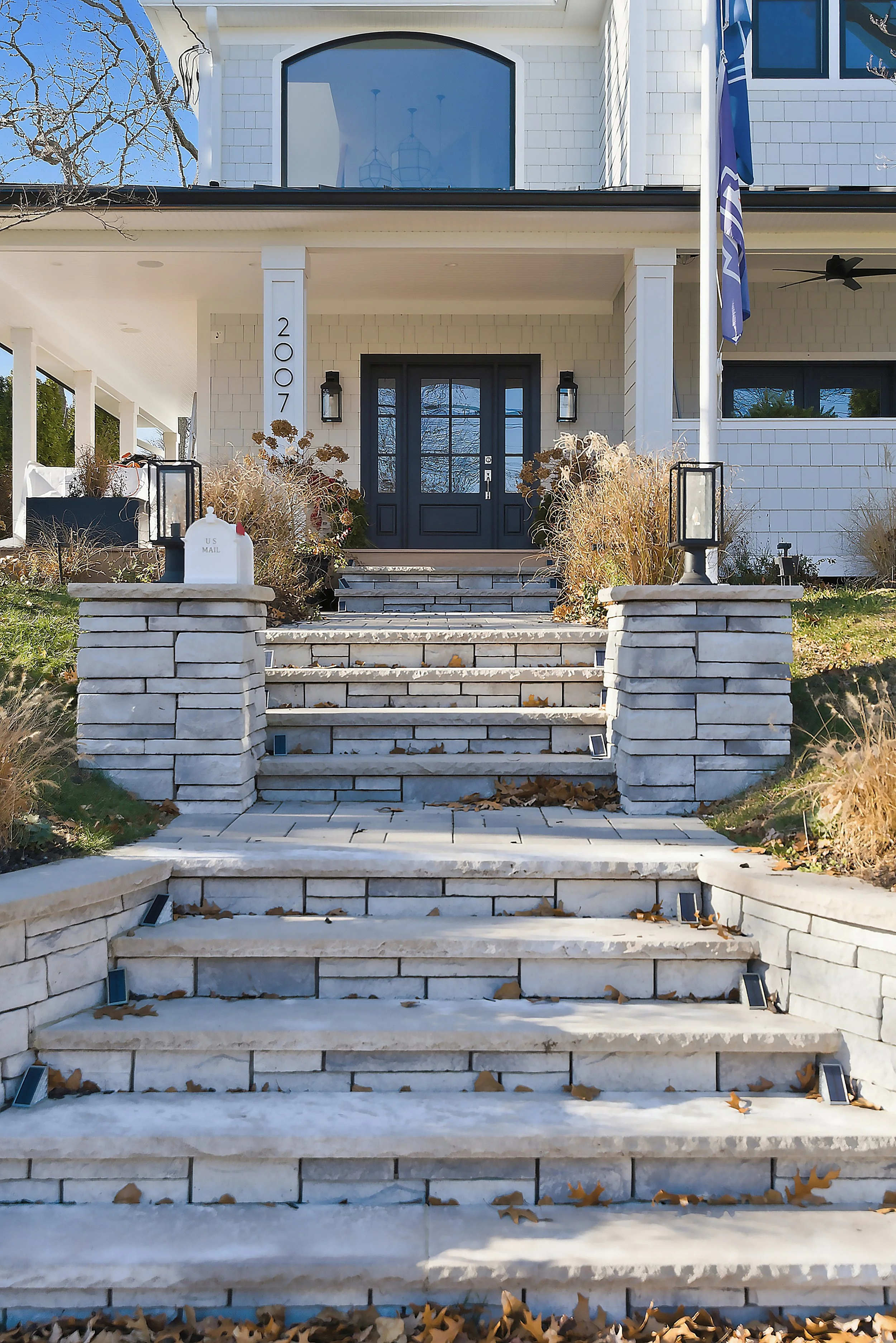 Front view of a white house with a dark front door, stone steps leading up to the entrance, and a mailbox on the left side of the steps. The house has white siding, black lantern-style lights, and a porch with two columns.