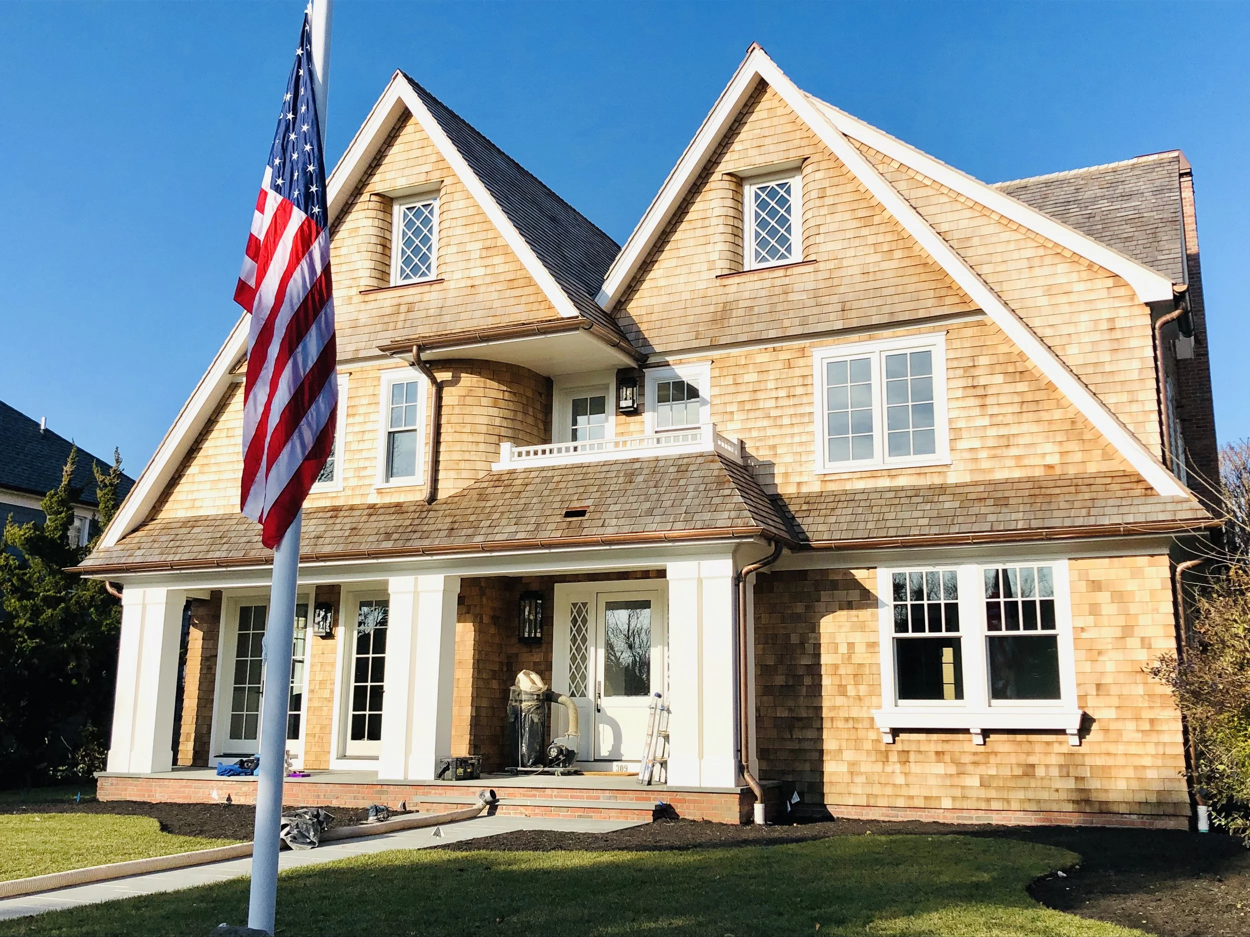 A large, multi-story house with tan shingle siding, white trim, and a front porch. An American flag is in the foreground. The sky is clear and blue.