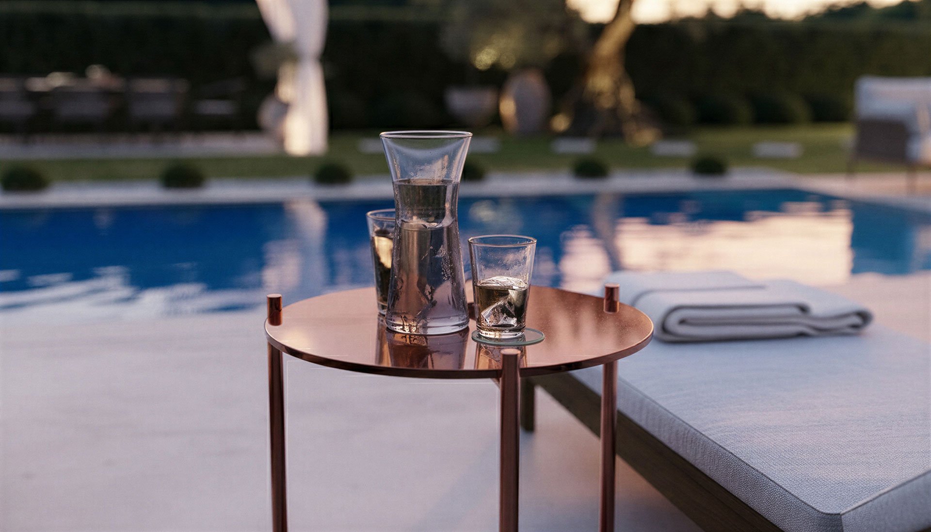 Photorealistic 3D render of glass pitcher and two glasses with ice and water on a small copper-colored table near a swimming pool.