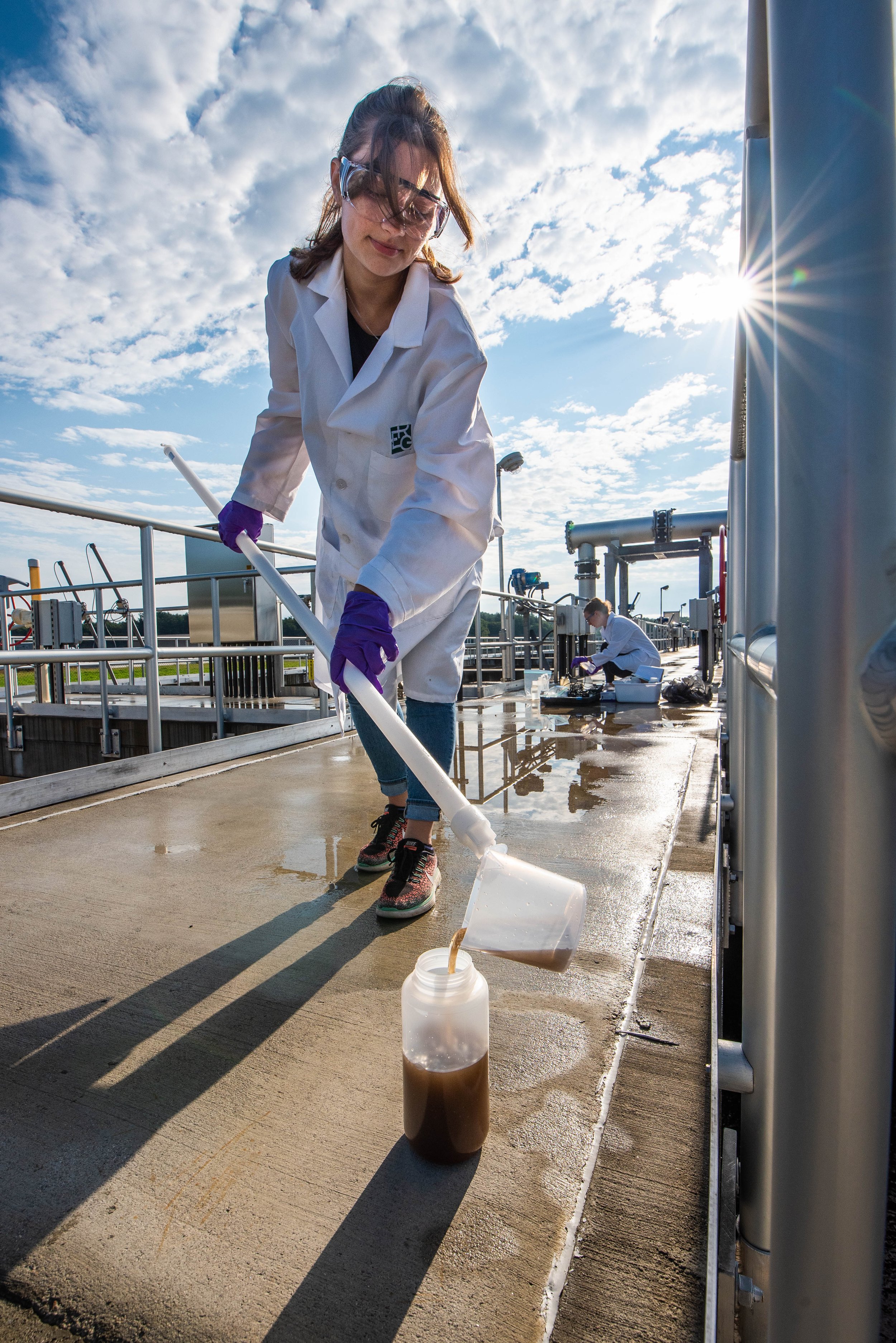 At a wastewater treatment facility on a concrete platform, a researcher in a white lab coat, safety goggles, and purple gloves pours a water sample out of a beaker on a pole into a bottle, while another researcher works behind her. S