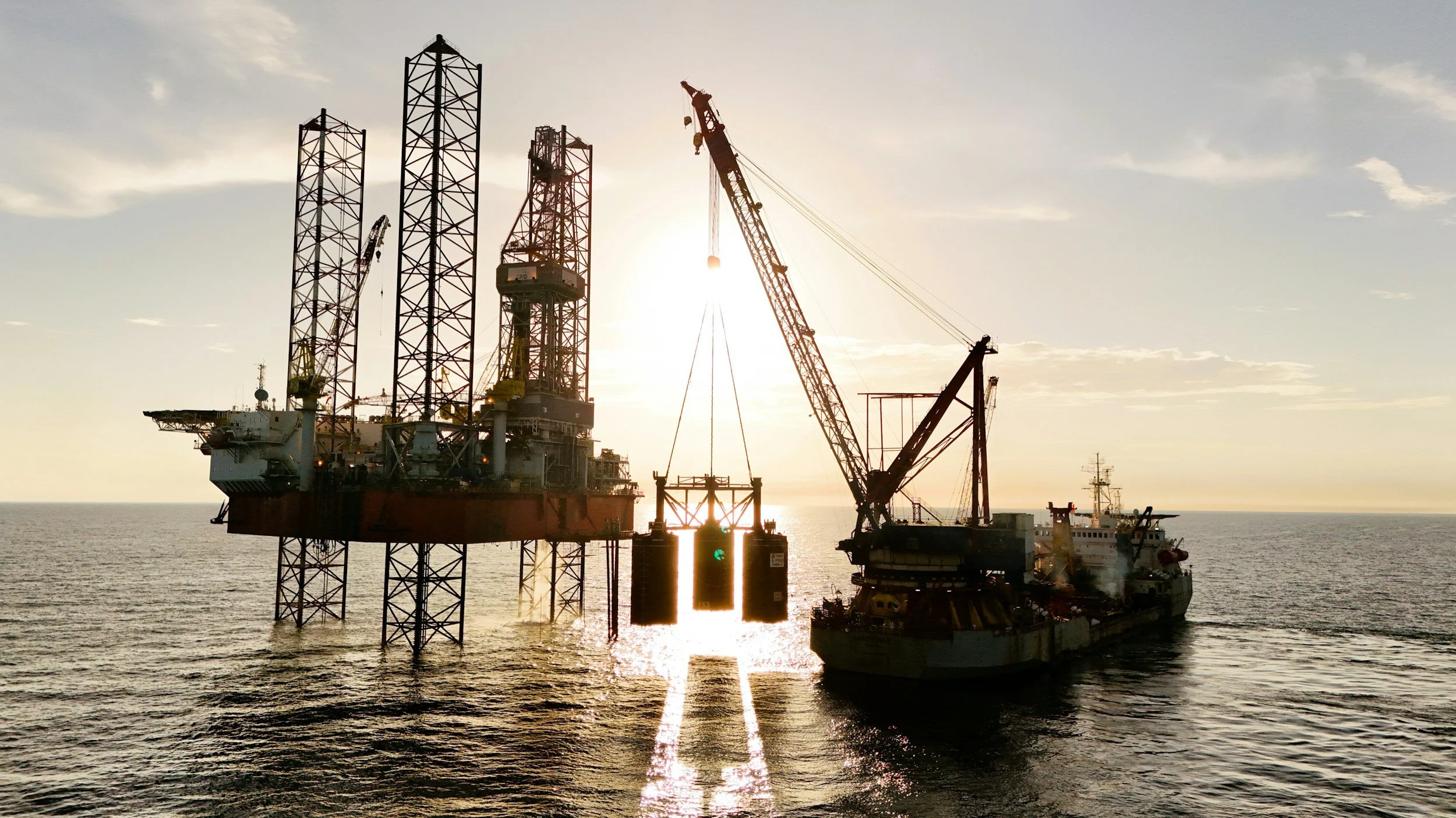 Offshore oil drilling platform with a crane barge nearby during sunset over the ocean.