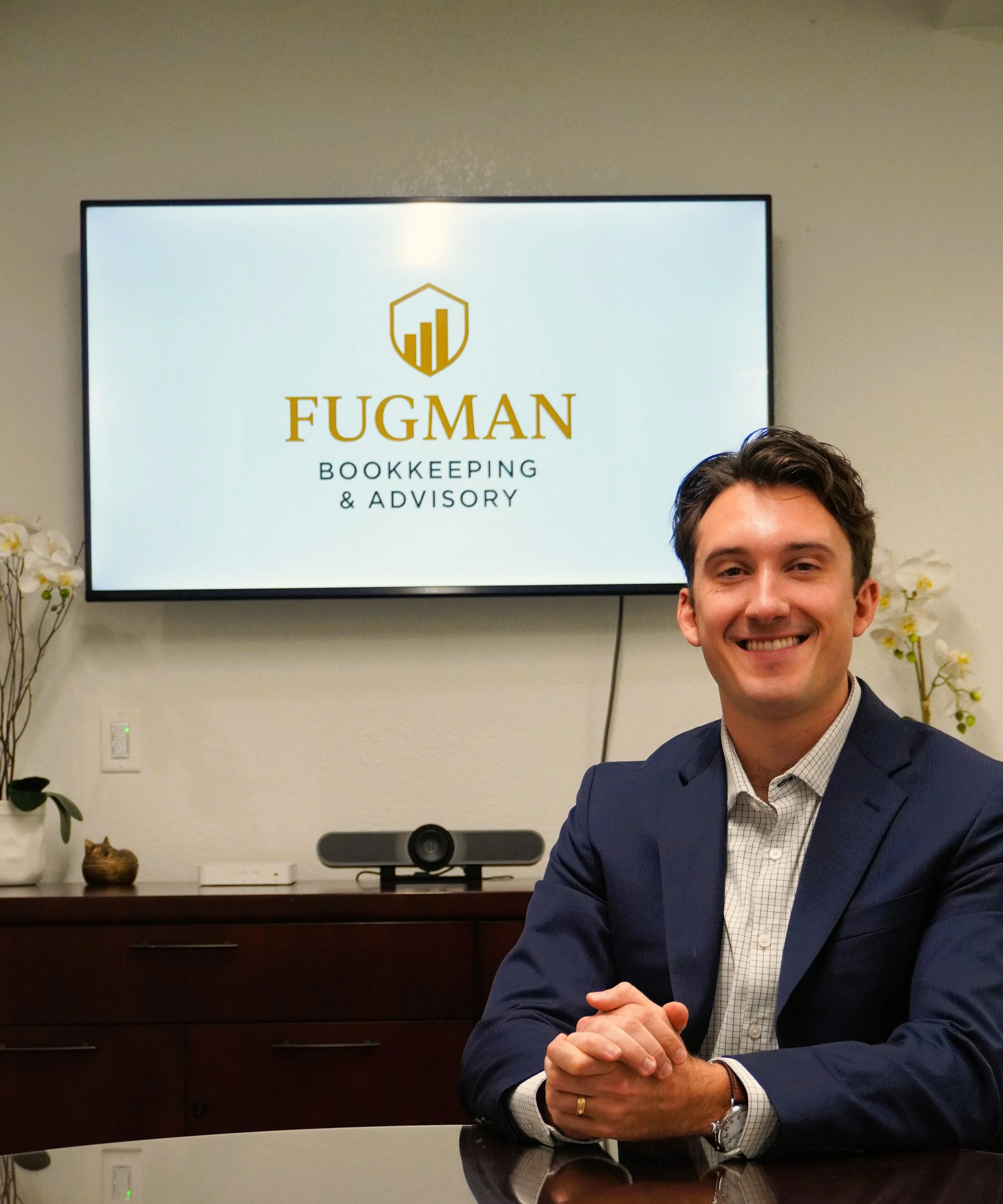 Smiling man in a dark suit sitting at a conference table with a logo for Fugman Bookkeeping & Advisory on a screen behind him.