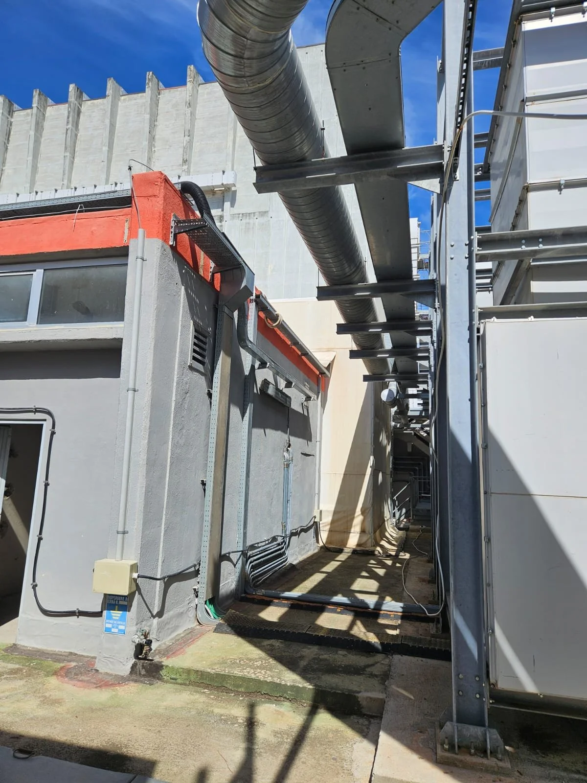 Immagine di un'area esterna di un edificio industriale con tubi metallici e strutture in metallo, ombreggiatura sul pavimento e un cielo sereno.