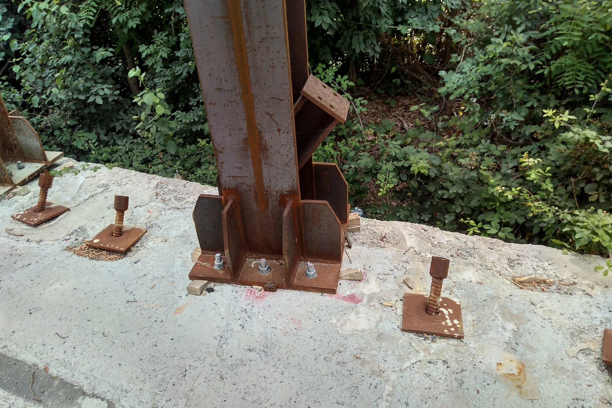 Struttura metallica di supporto tra due pilastri di cemento, con bulloni di fissaggio e piante verdi sullo sfondo.