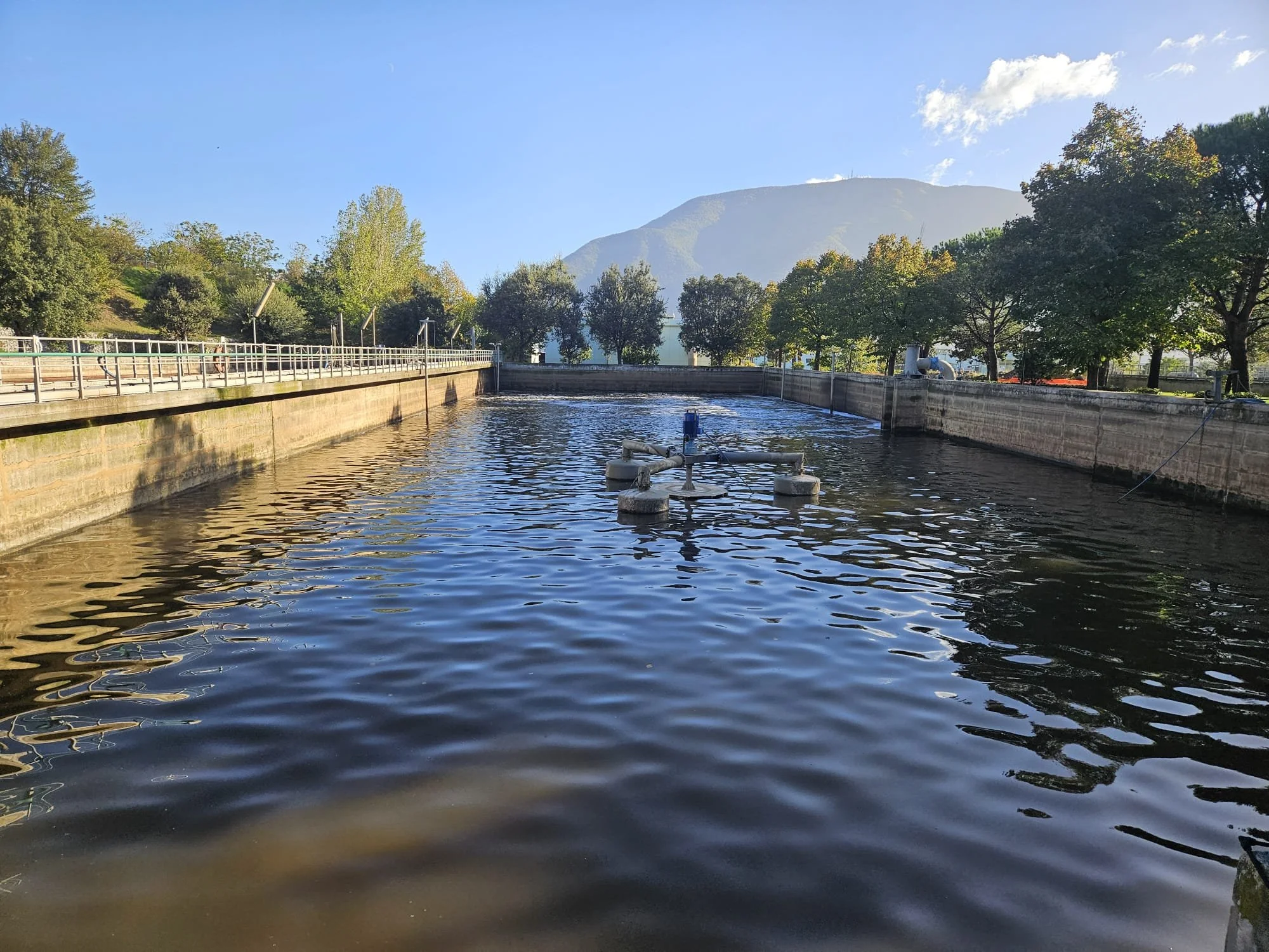 Corso d'acqua con apparecchiature di pompaggio, recinzioni lungo le sponde e albero sullo sfondo, montagne e cielo sereno.