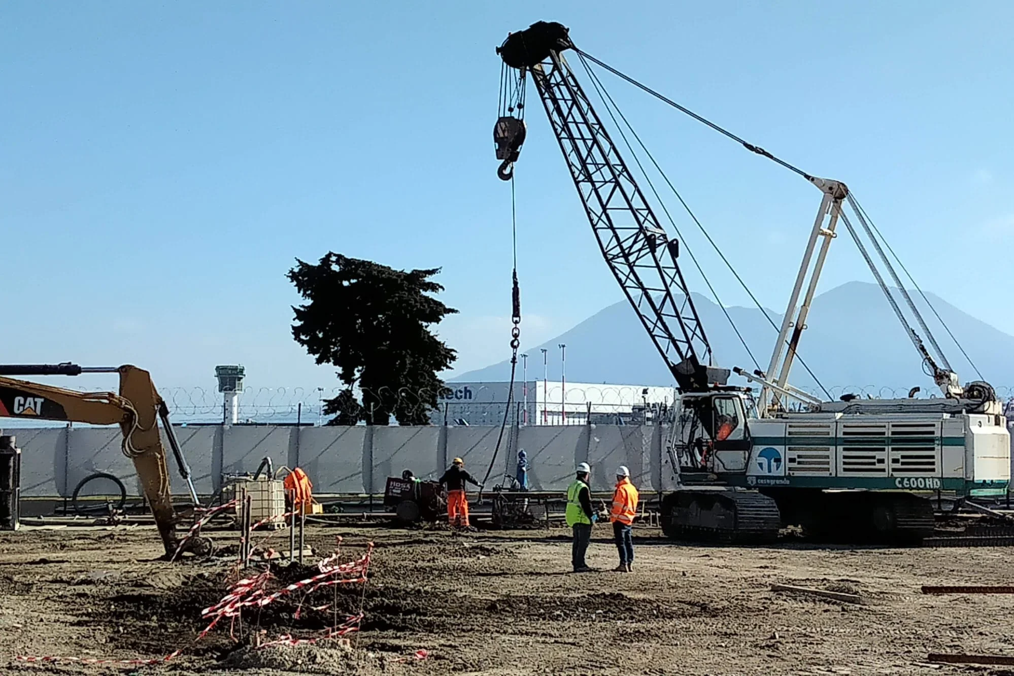 Operai sul cantiere con grandi gru e attrezzi da costruzione, recinzione e montagne sullo sfondo.