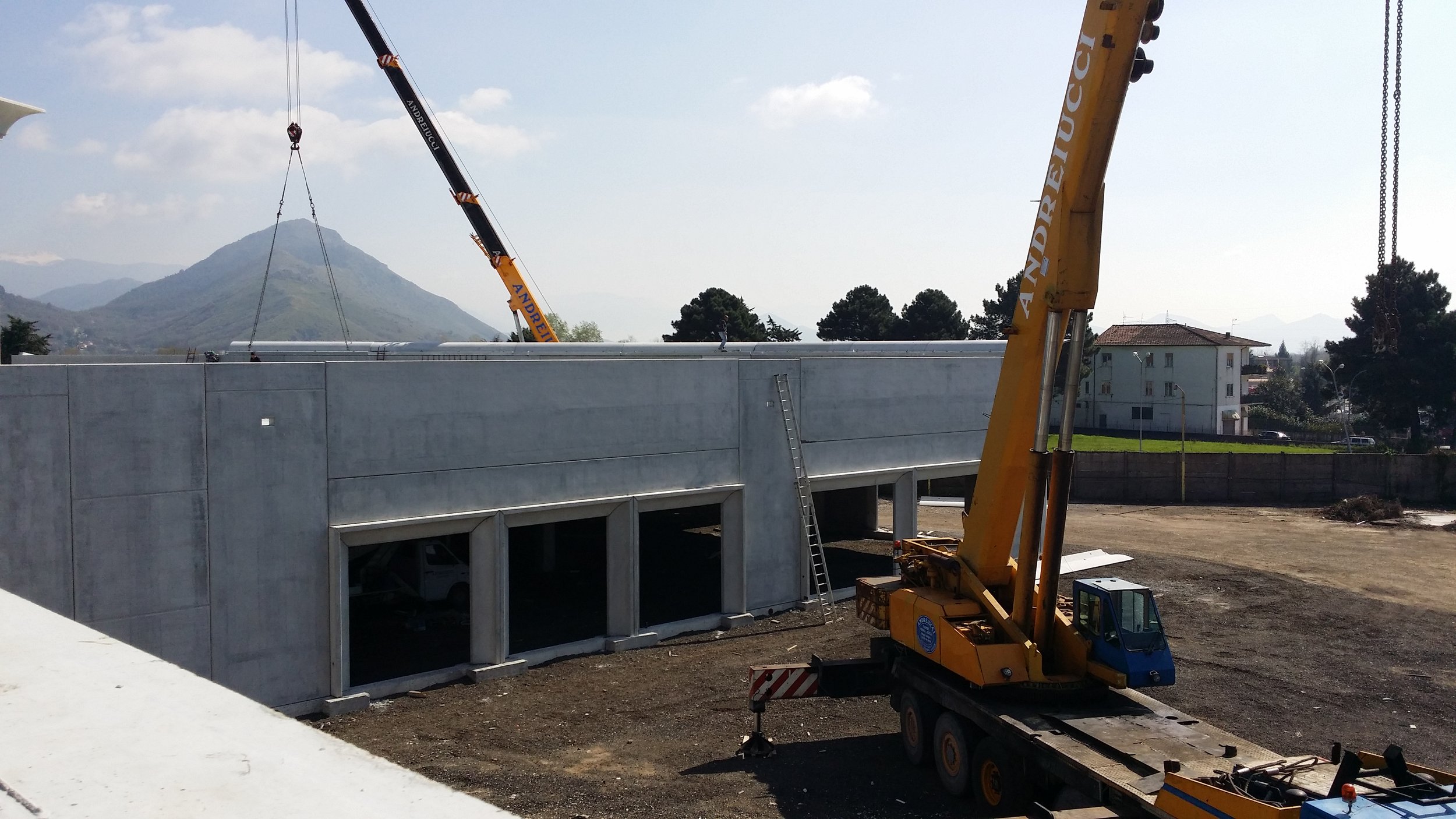 Cantiere edile con gru gialla che solleva strutture sopra un edificio in costruzione, con montagne sullo sfondo.