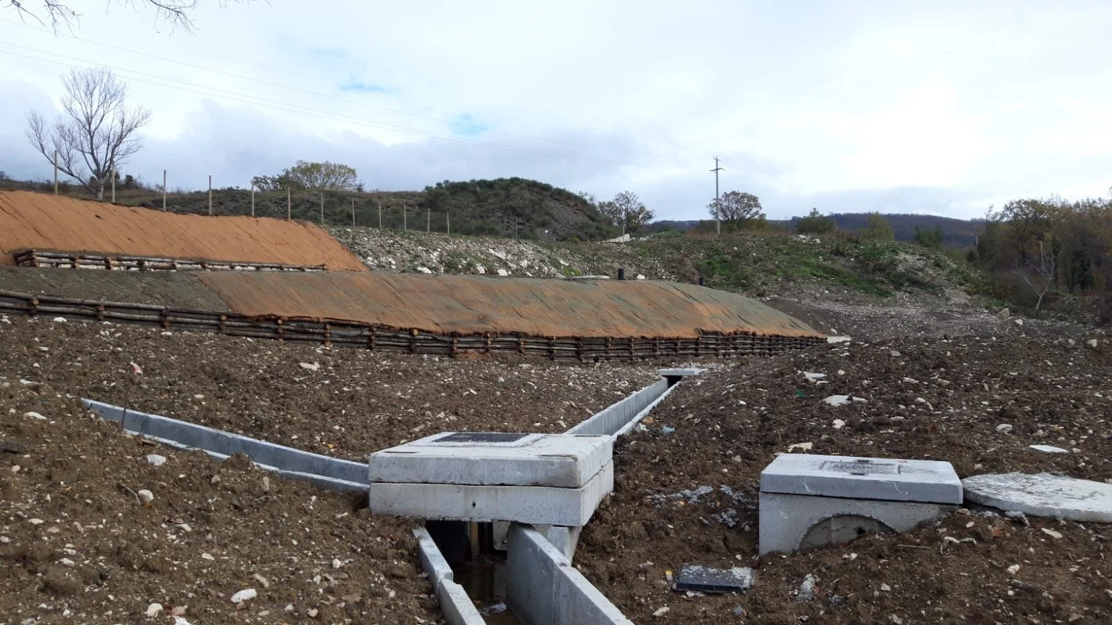 Un cantiere con fondamenta e tubazioni in cemento in un'area all'aperto, con un terreno in terra smossa e alcune colline in lontananza.