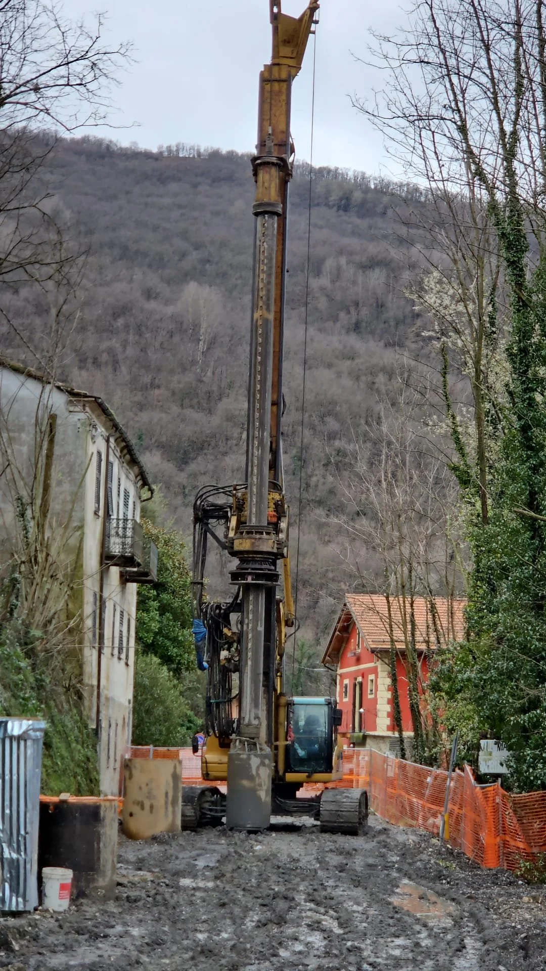 Macchina escavatore in un cantiere stradale circondato da recinzioni arancioni, con case e montagne sullo sfondo.