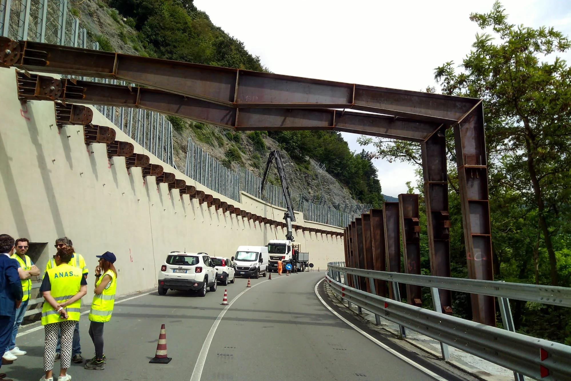 Lavori in corso sulla strada con veicoli e persone in gilet di sicurezza gialli, con una grande struttura metallica sopra la strada e un ponteggio di ferro
