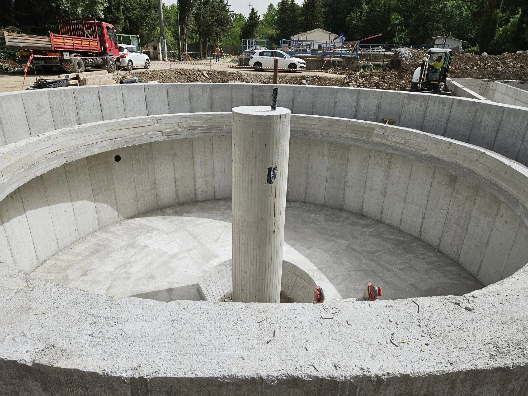 Struttura in cemento in costruzione, che sembra essere parte di un impianto idraulico o di un pozzetto, con un grande foro centrale e una colonna verticale nel mezzo.