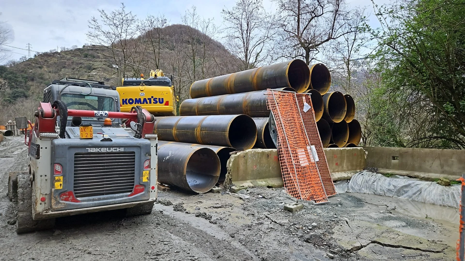 Cantiere stradale con tubi di grandi dimensioni e attrezzature da lavoro, tra cui una macchina gialla Komatsu e una macchina rossa Takeuchi.