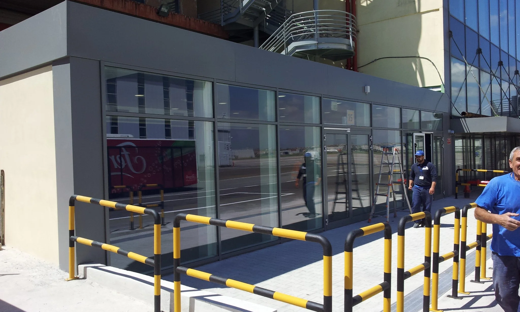 Ingresso di un edificio con pareti di vetro, accesso pedonale con barriera gialla e nera, persone intorno, alcuni con tenuta da lavoro, l'edificio ha una facciata moderna con finestre a specchio.