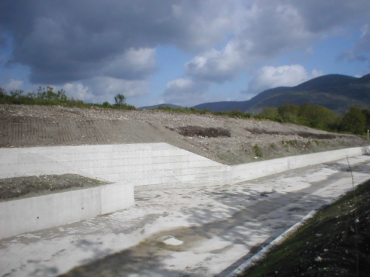Costruzione di un muro di contenimento in cemento lungo una strada in una zona rurale con montagne sullo sfondo e un cielo nuvoloso.