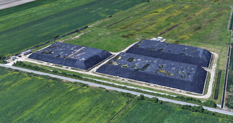 Immagine aerea di edifici industriali con tetti danneggiati e piante cresciute sul tetto, circondati da campi verdi.