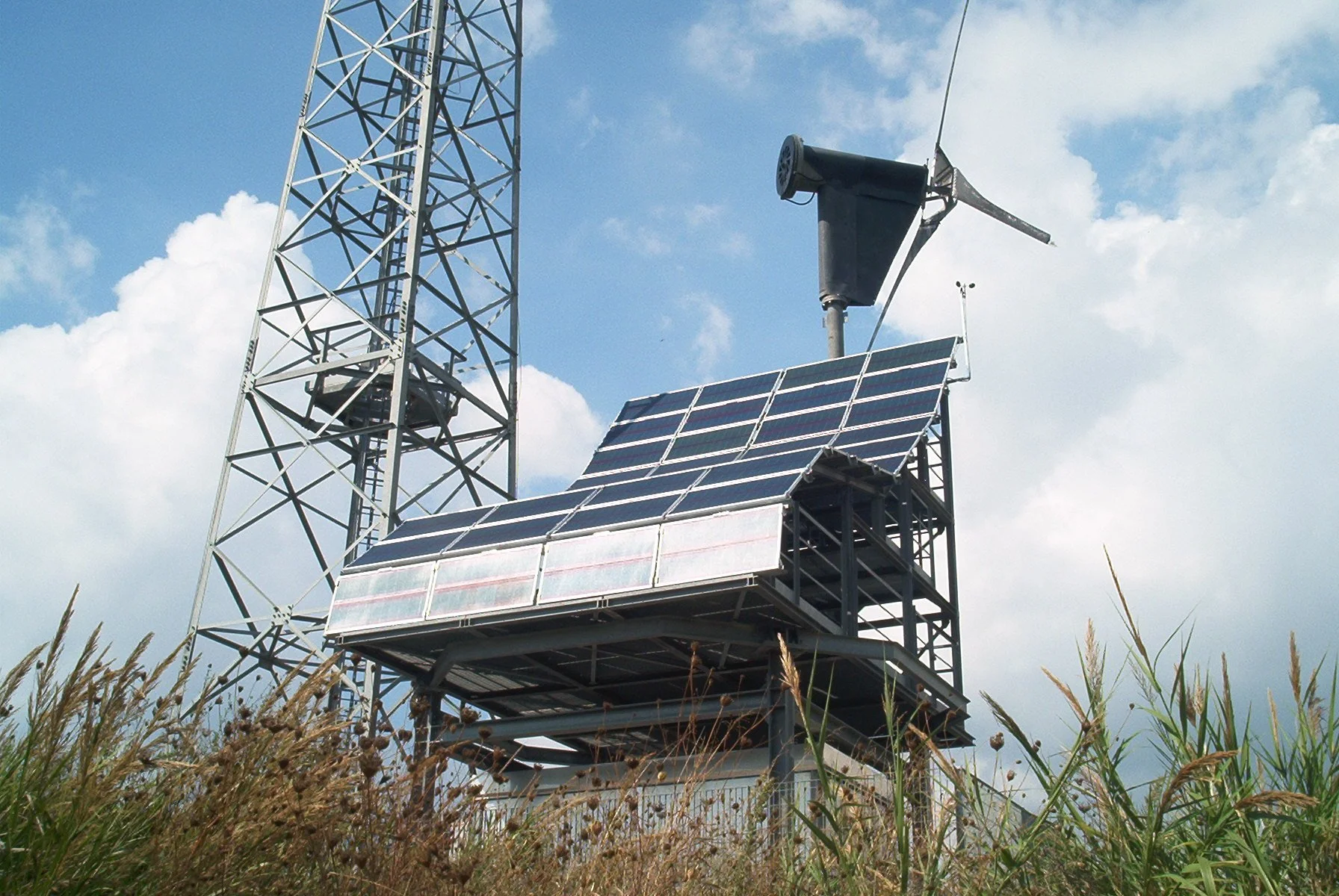 Pannelli solari e apparecchiature meteorologiche su una torre in un campo con erba, sotto un cielo nuvoloso.