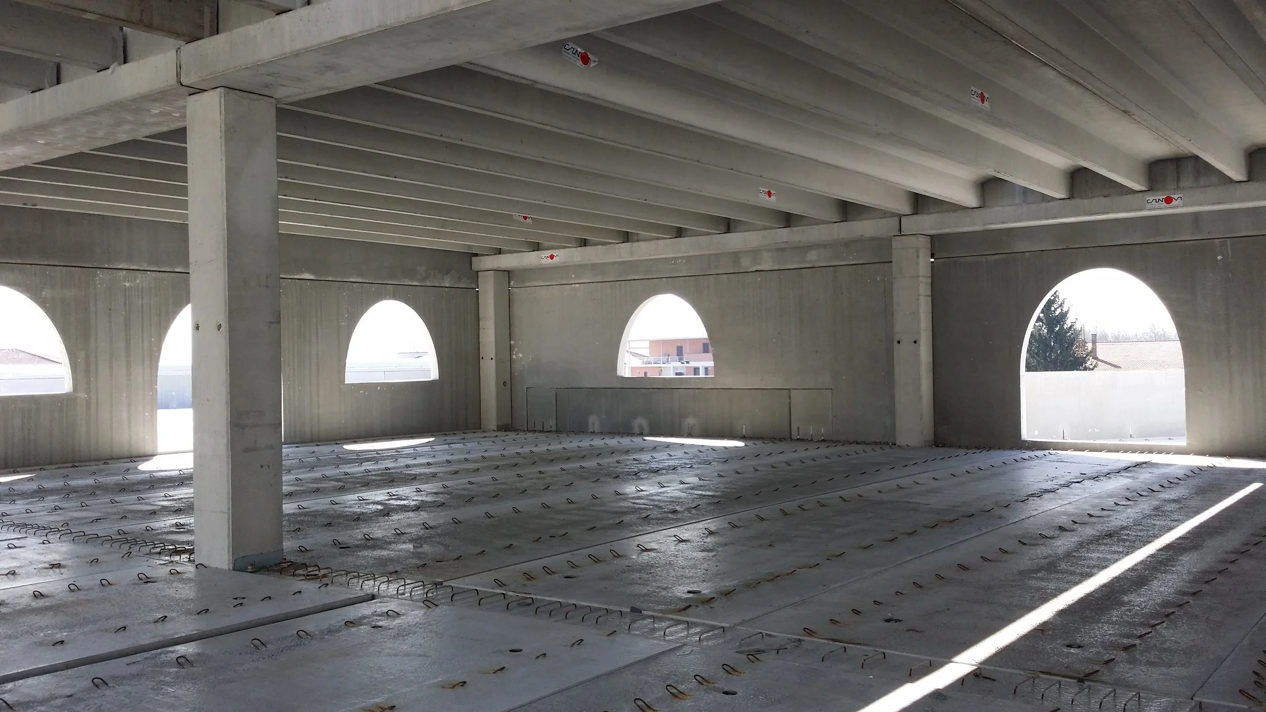 Interno di un edificio in costruzione con muri e soffitto in cemento, apertura ad archi lungo le pareti e un pavimento ancora in fase di finitura con elementi di ancoraggio.