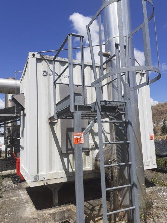 Struttura metallica con scala e gabbia protettiva, parte di un impianto industriale all'aperto contro cielo sereno.