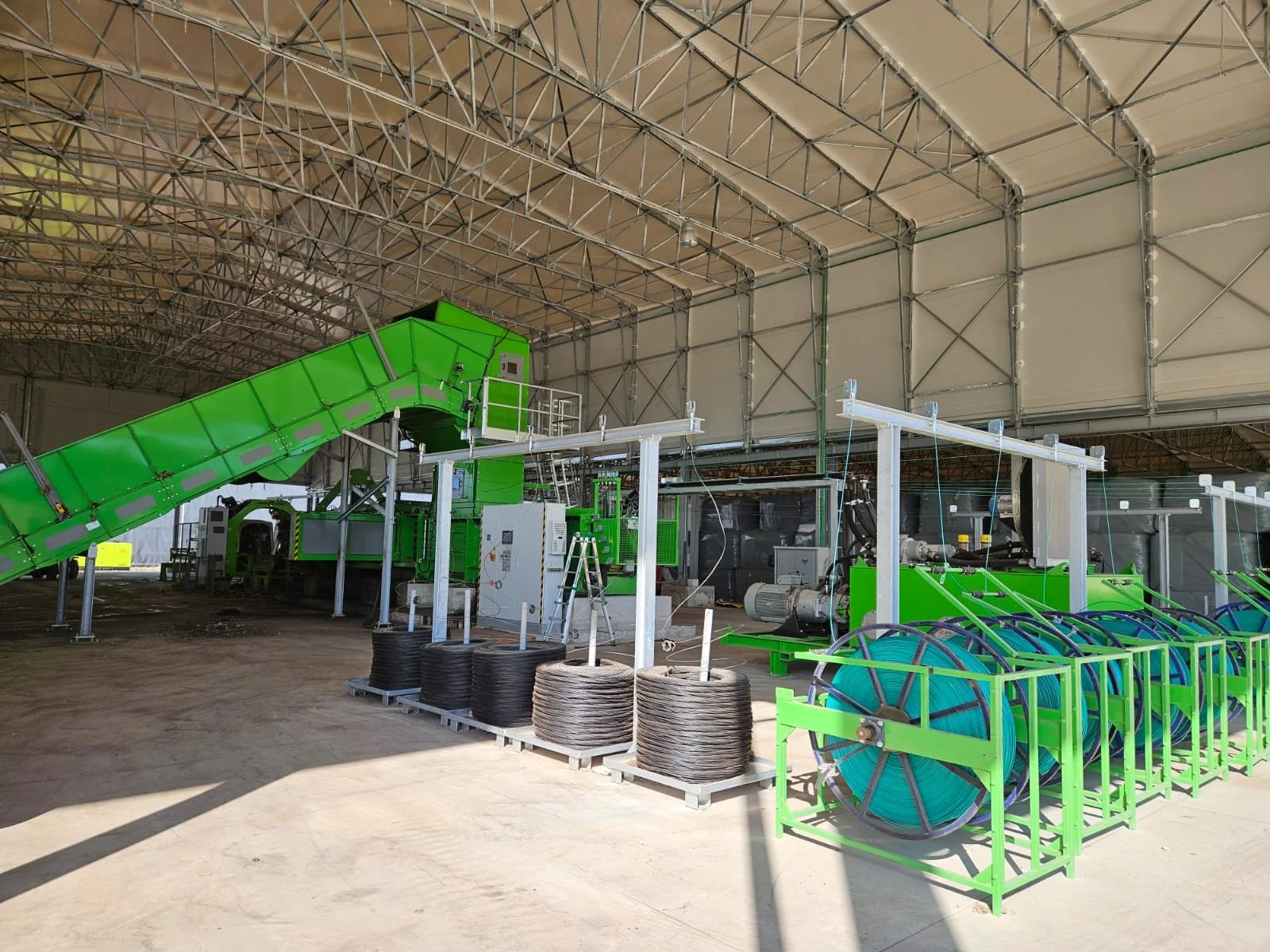 Interno di un grande magazzino con attrezzature industriali, tubi di gomma, bobine di cavi e una macchina verde, sotto una copertura con struttura in metallo.