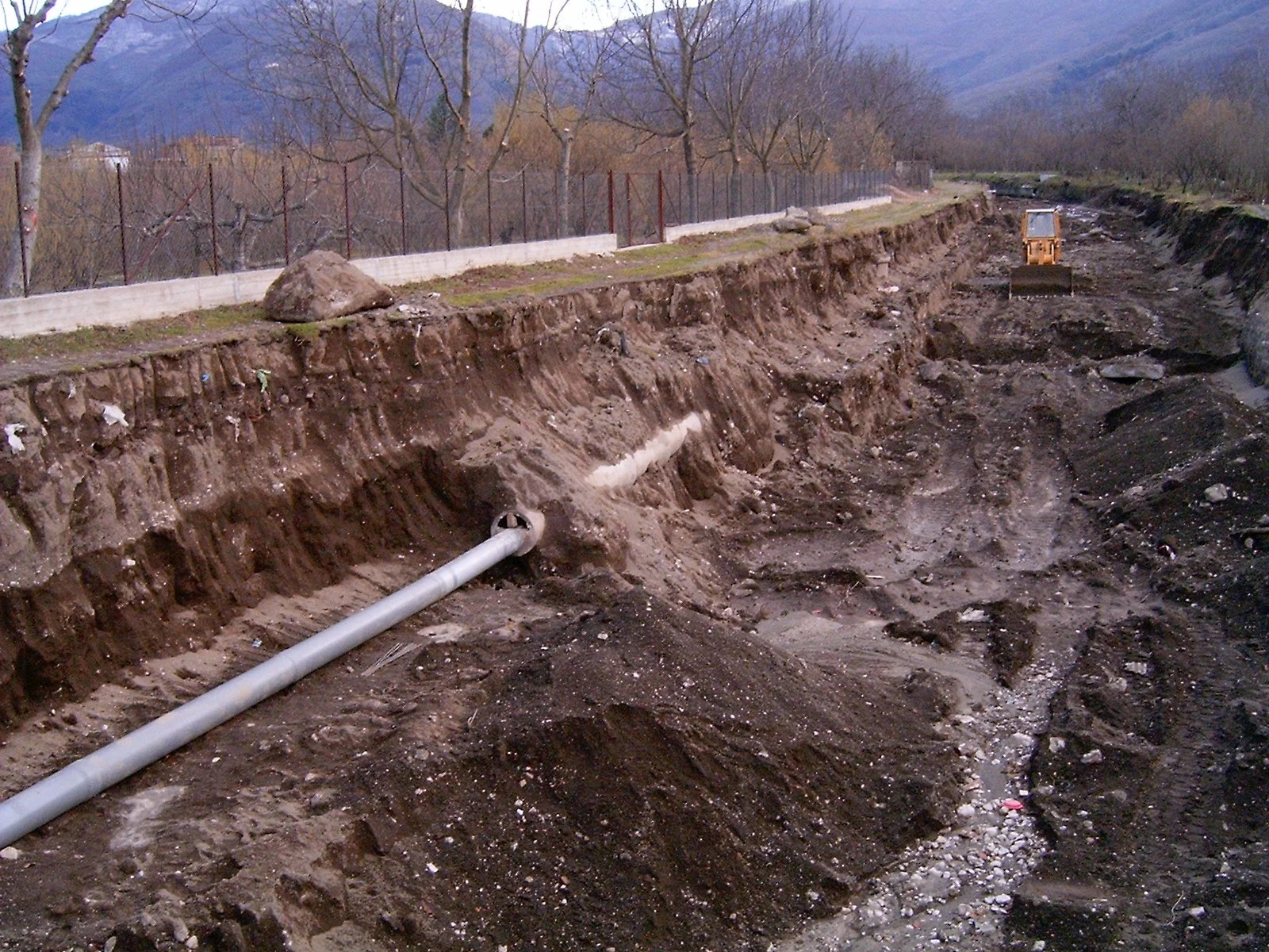 Lavori di scavo e posa di tubature in un terreno a scavi, con una piccola macchina escavatrice in fondo.