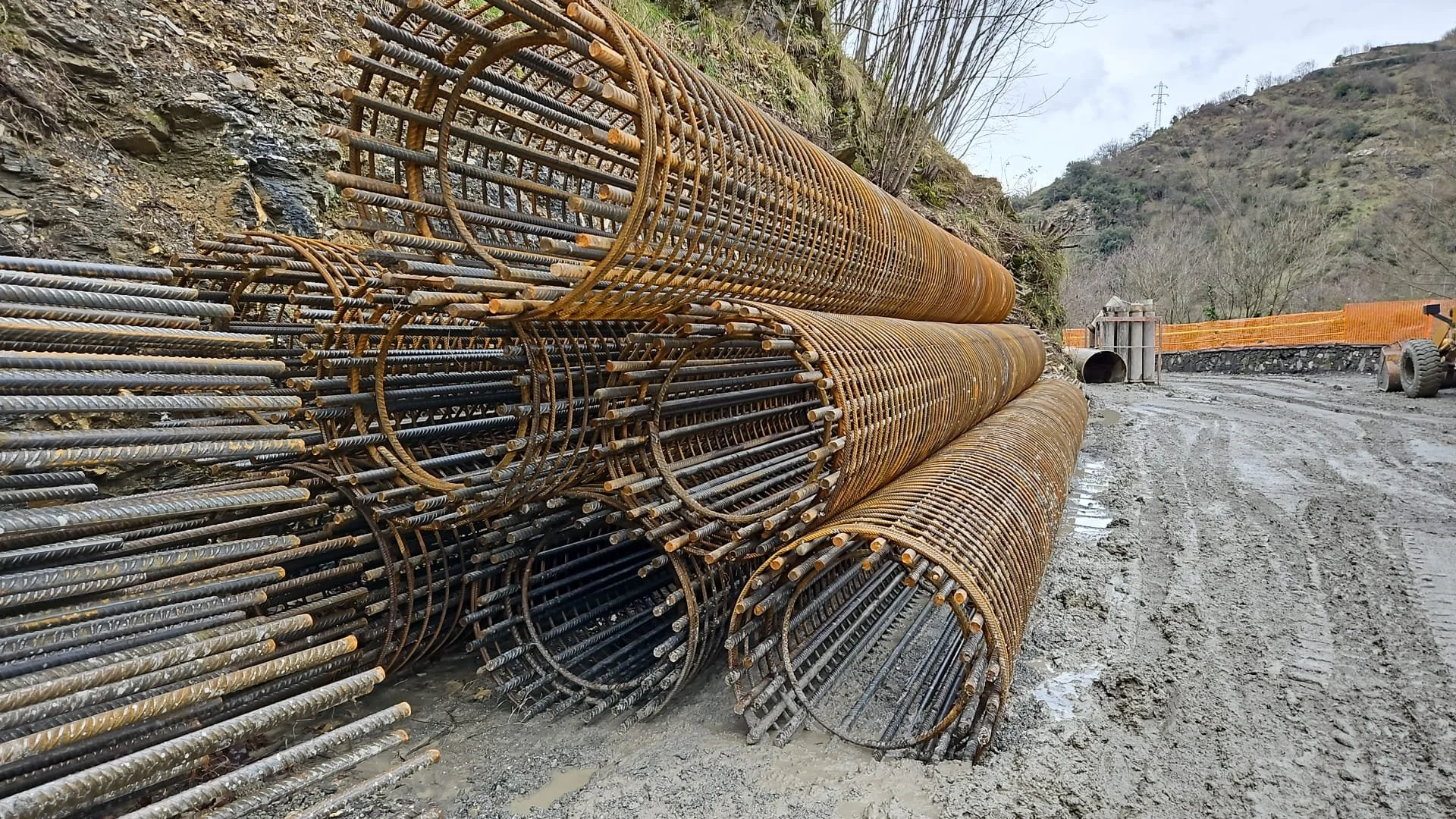 Pili di barre d'acciaio arrugginite stoccati su un cantiere stradale, con terriccio, montagna e attrezzature di costruzione in background.