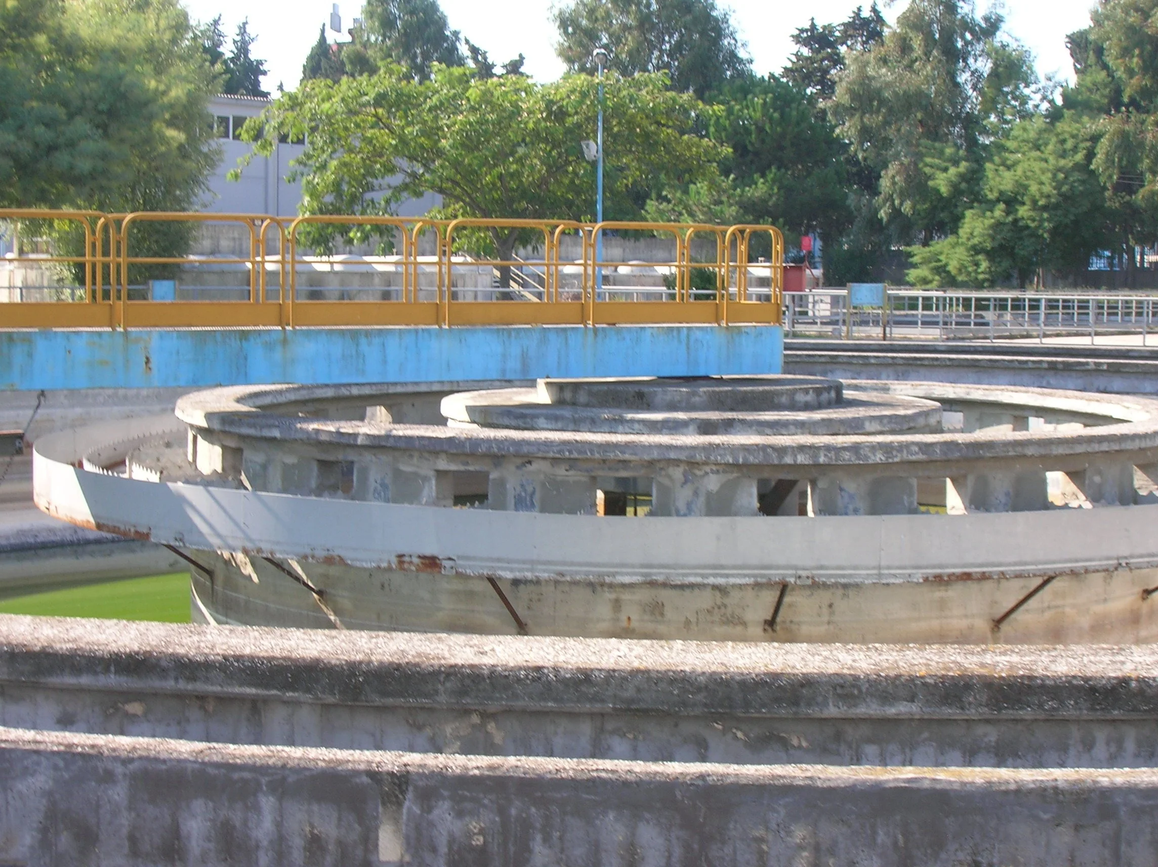 struttura di un impianto di trattamento delle acque con vasche di cemento e ringhiere gialle, in un'area all'aperto con alberi e edifici sullo sfondo